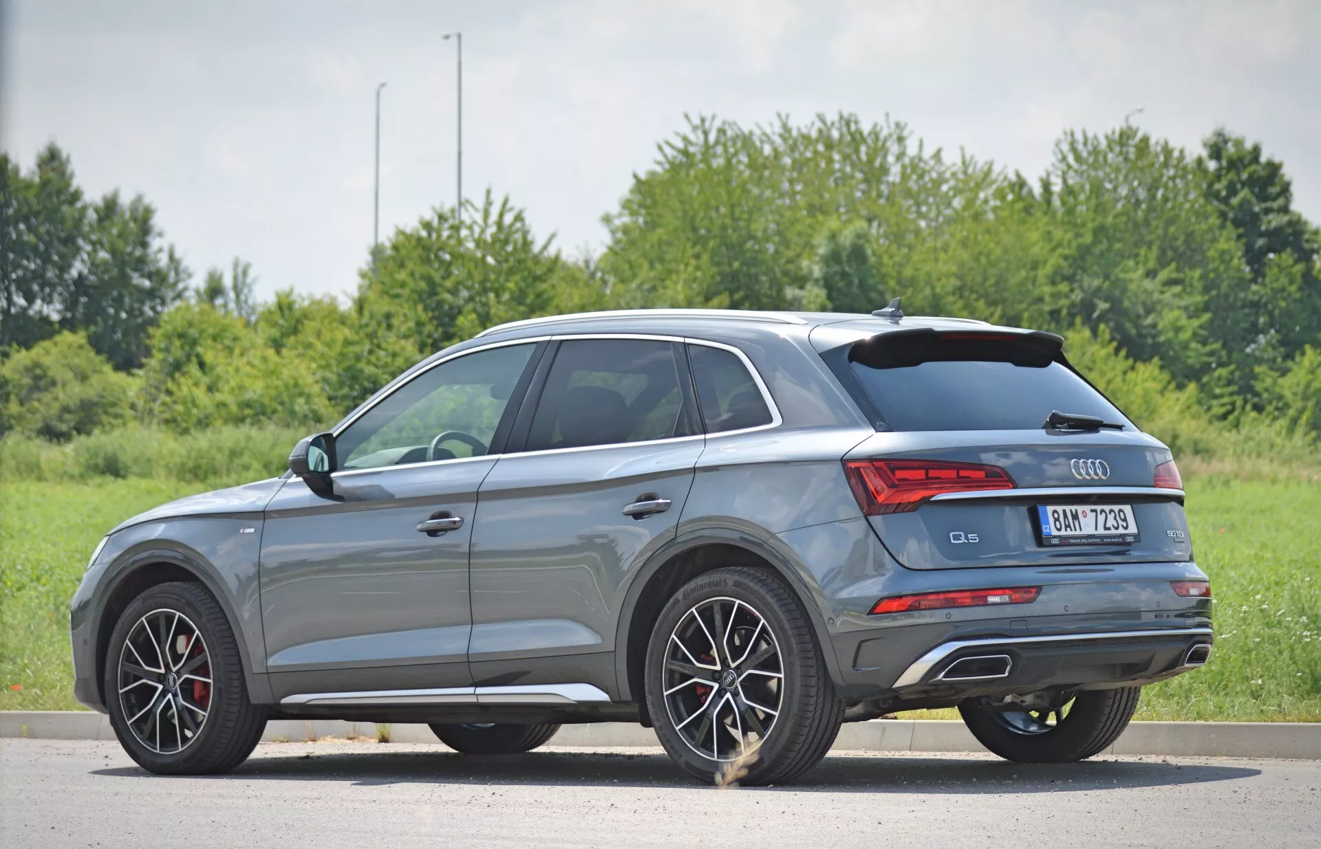 Šedé SUV Audi Q5 Sportback z modelového roku 2020 je zobrazené z trojštvrťového zadného pohľadu, ktorý zdôrazňuje elegantné línie karosérie, viaclúčové zliatinové kolesá, LED zadné svetlá, dvojité výfukové koncovky a klesajúcu zadnú strešnú líniu. Auto je zaparkované na rovnej ploche s prirodzeným pozadím trávy a stromov, pričom prirodzené denné svetlo zvýrazňuje športové dizajnové prvky a kompaktný vzhľad stredne veľkého SUV.
