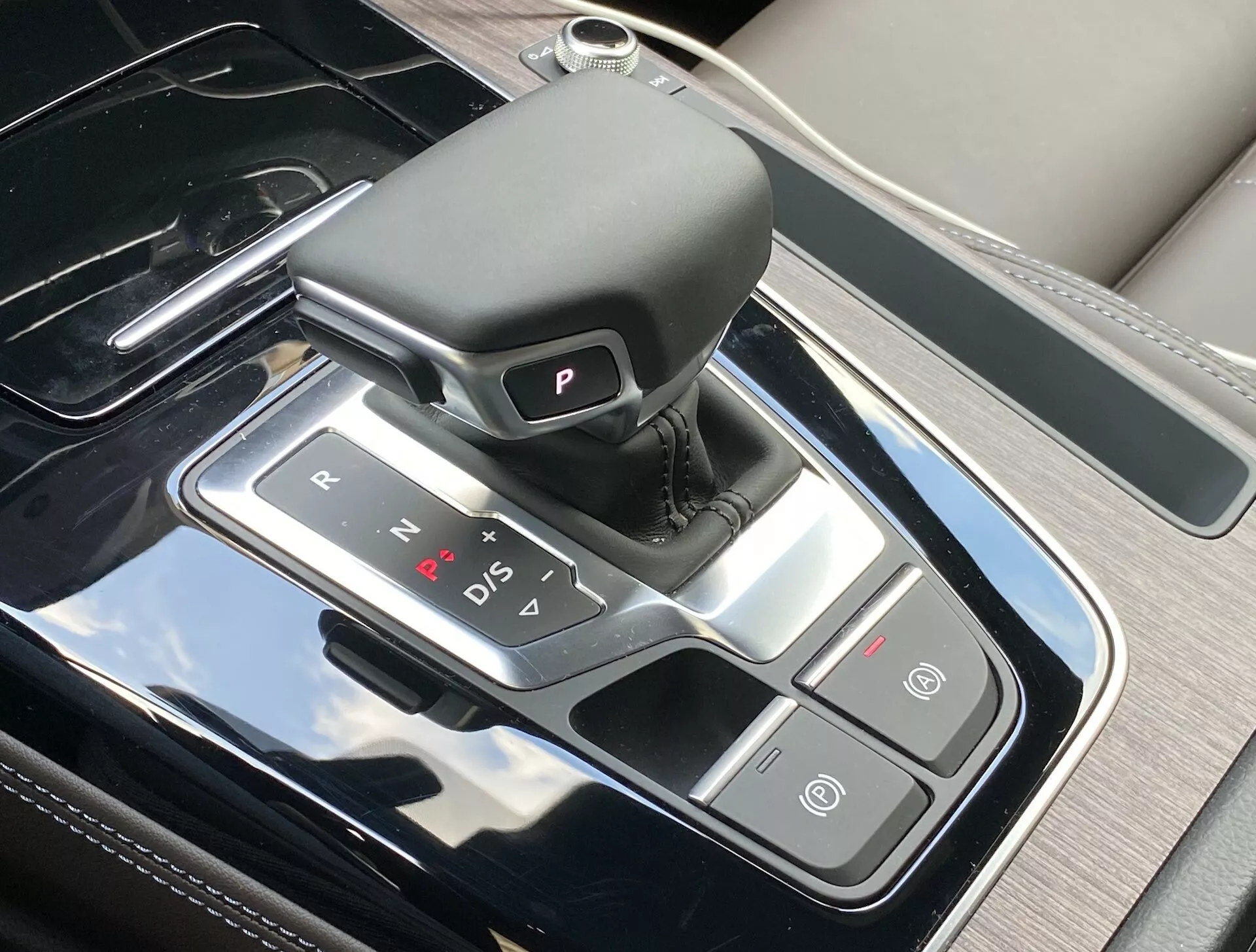 Pohľad zhora na stredovú konzolu Audi Q5 Sportback 2020, zvýrazňujúci prepracovanú oblasť radiacej páky s koženým automatickým voličom, osvetlenými indikátormi prevodových stupňov a priliehajúcimi ovládacími tlačidlami pre automatickú parkovaciu brzdu a funkciu Auto Start/Stop. Konzola má lesklý čierny povrch so strieborným lemovaním, čo odráža luxusný interiérový dizajn a modernú technológiu.