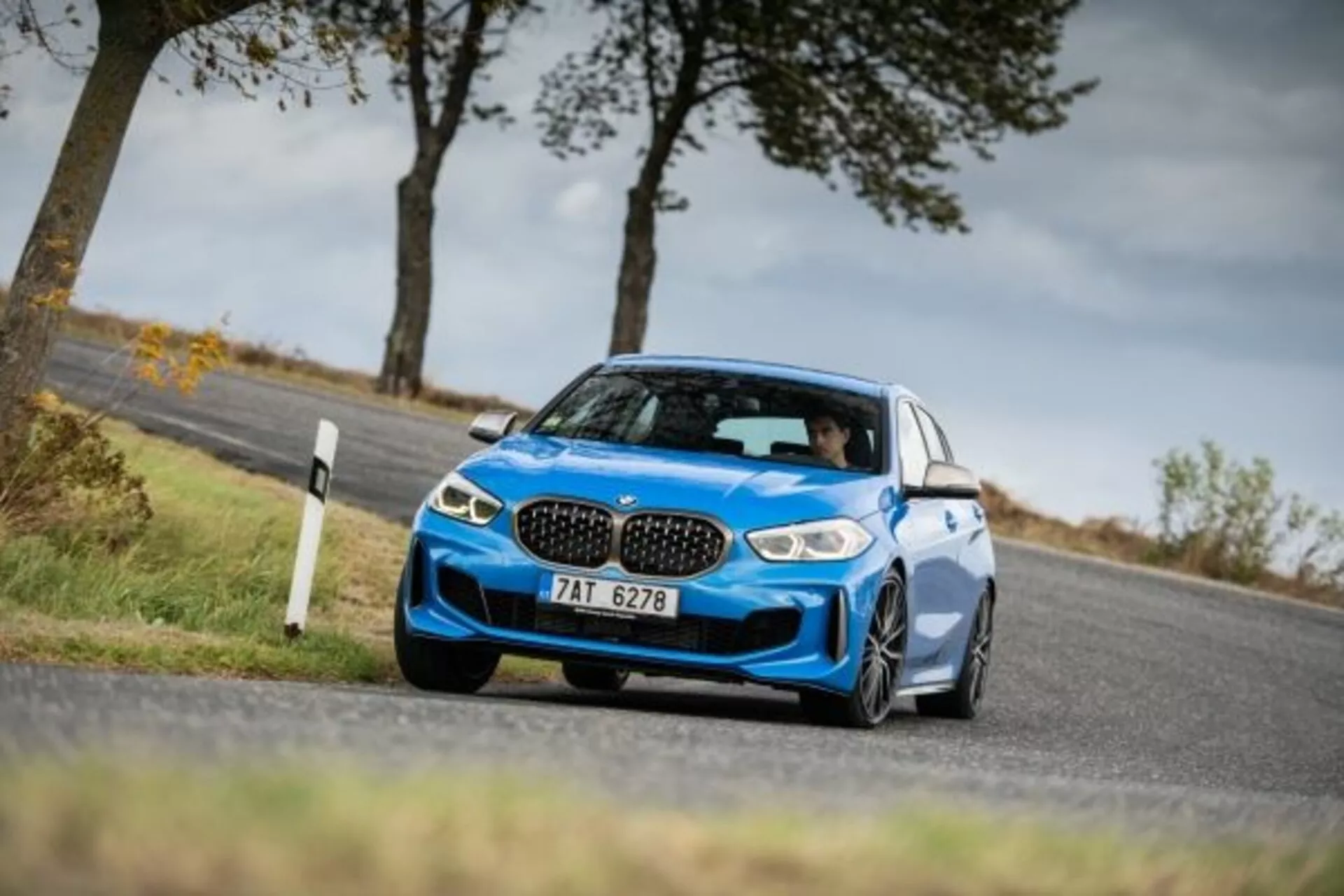 Dynamická fotografia z nízkeho uhla moderného modrého športového sedanu BMW radu 1 M Performance z roku 2019 v pohybe na vidieckej ceste, zobrazujúca jeho moderný dizajn, charakteristickú obličkovitú masku, agresívne LED svetlomety, prepracovaný predný nárazník so vzduchovými vstupmi a zliatinové kolesá