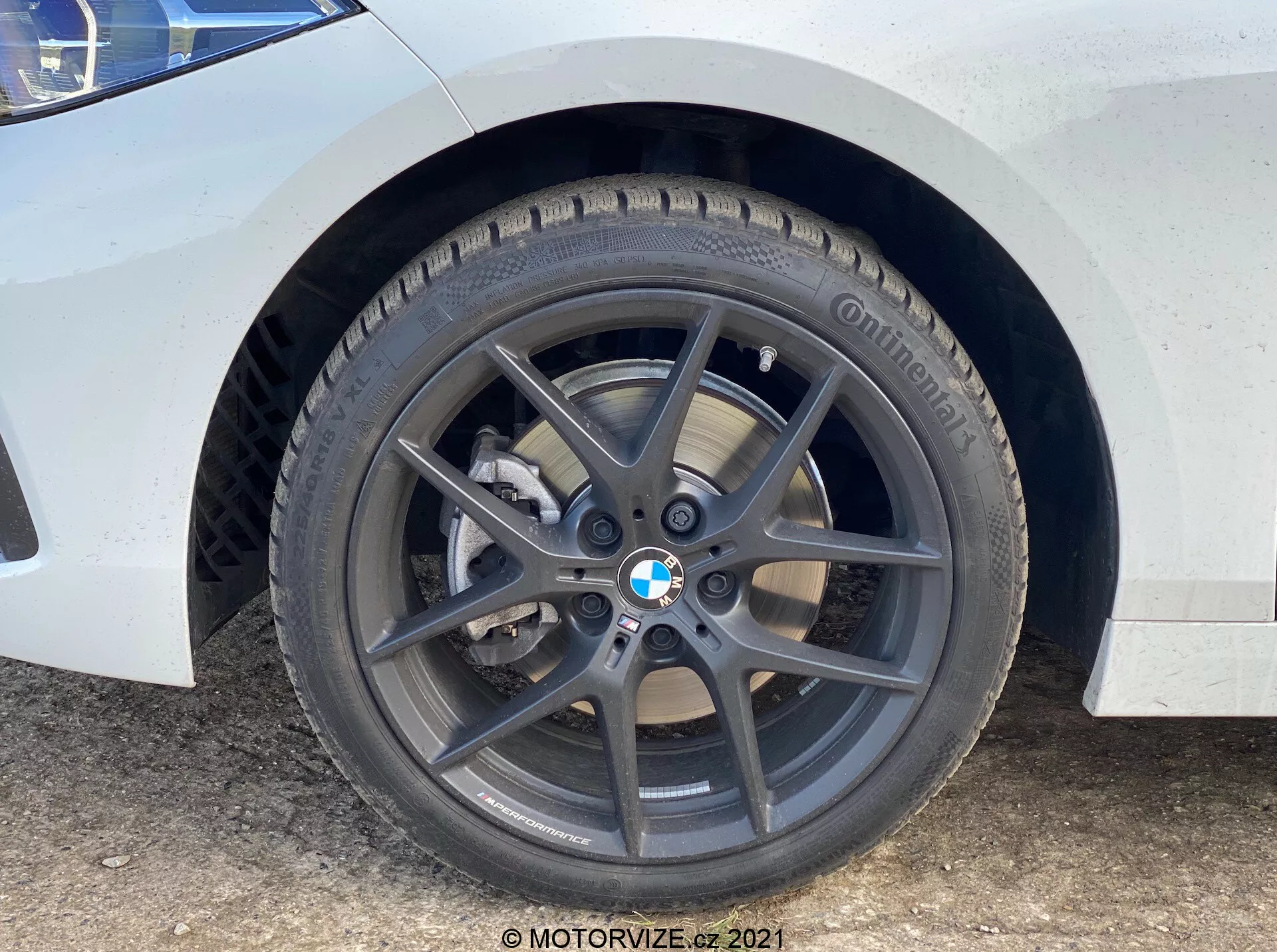 Detailný záber na predné koleso a blatník BMW 1er-Reihe 2019 (BMW-116i). Perspektíva z úrovne zeme zdôrazňuje tmavosivý päťzväzkový hliníkový disk s logom BMW, značkovú pneumatiku Continental s viditeľným dezénom, ventilovaný brzdový kotúč a strieborný jednopiestny brzdový strmeň. Biela karoséria s nečistotami a škvrnami vody indikuje stav používania.
