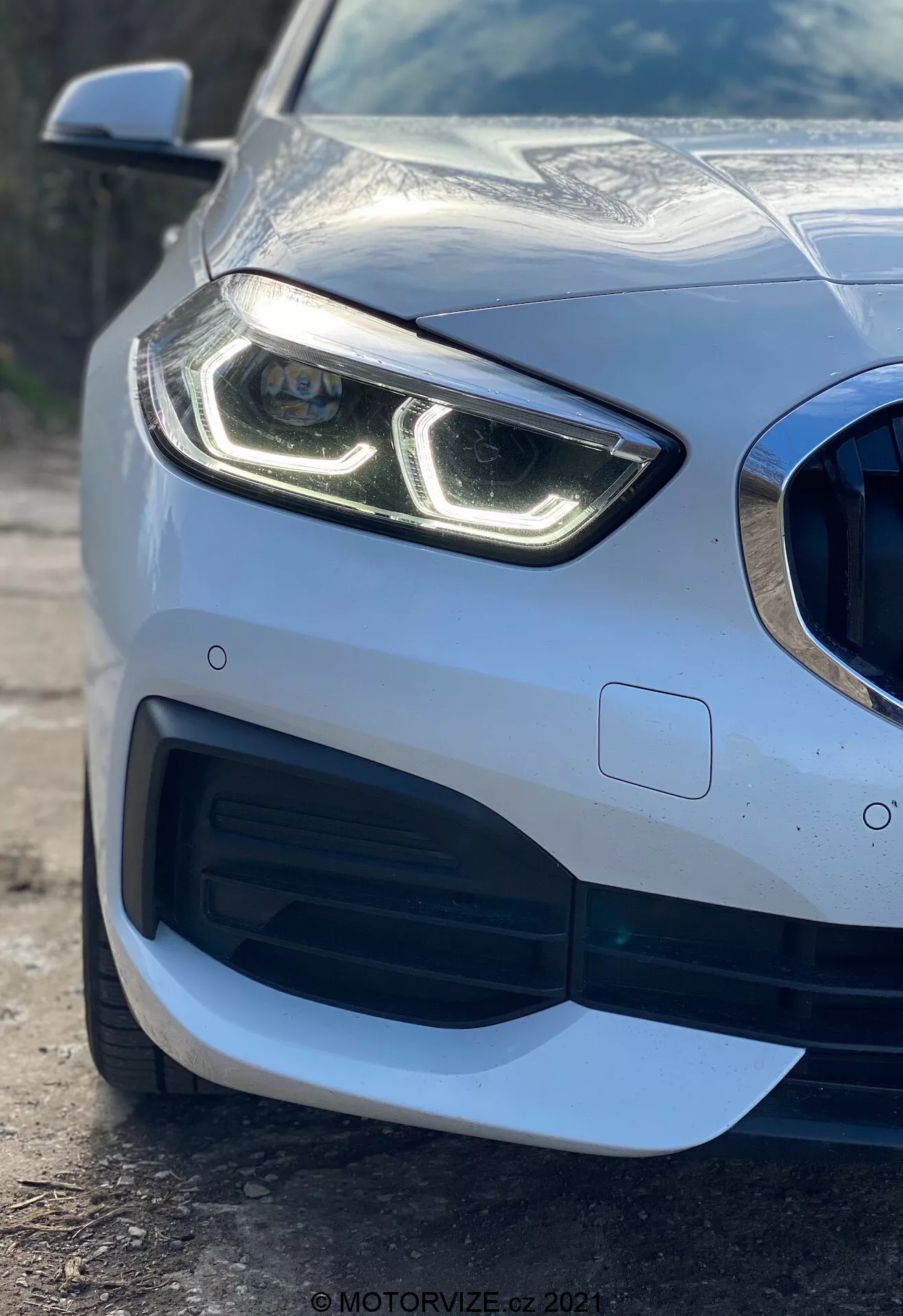 Detailný pohľad na prednú ľavú časť vozidla BMW radu 1 2019 (BMW 116i) s dôrazom na moderný svetlomet s výraznými LED dennými svetlami, mriežku chladiča vo včelím vzore s veľkým prívodom vzduchu, sofistikovaný aerodynamický dizajn predného nárazníka a parkovací senzor integrovaný v nárazníku, ukazujúci prémiový metalický lak a lesklý povrch karosérie.