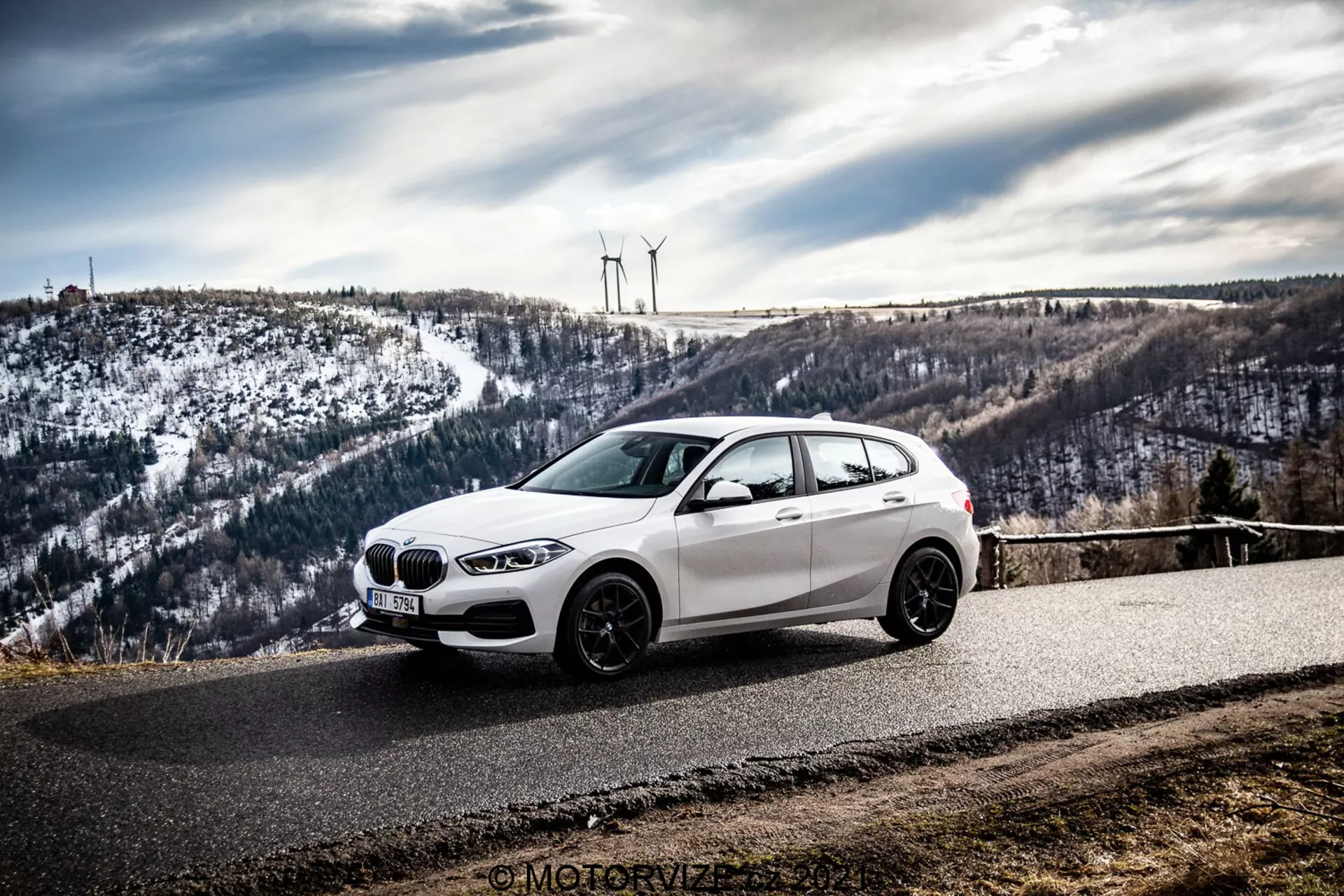 Biele hatchback BMW, pravdepodobne BMW 1er-Reihe 2019 (BMW 116i), je zaparkované na zakrivenej asfaltovej ceste obklopenej kopcovitou, zasneženou krajinou s veternými turbínami. Fotografia, urobená z bočno-predného uhla, zvýrazňuje prednú mrežu, svetlomety, aerodynamické línie a zliatinové kolesá auta pod zamračeným nebom.
