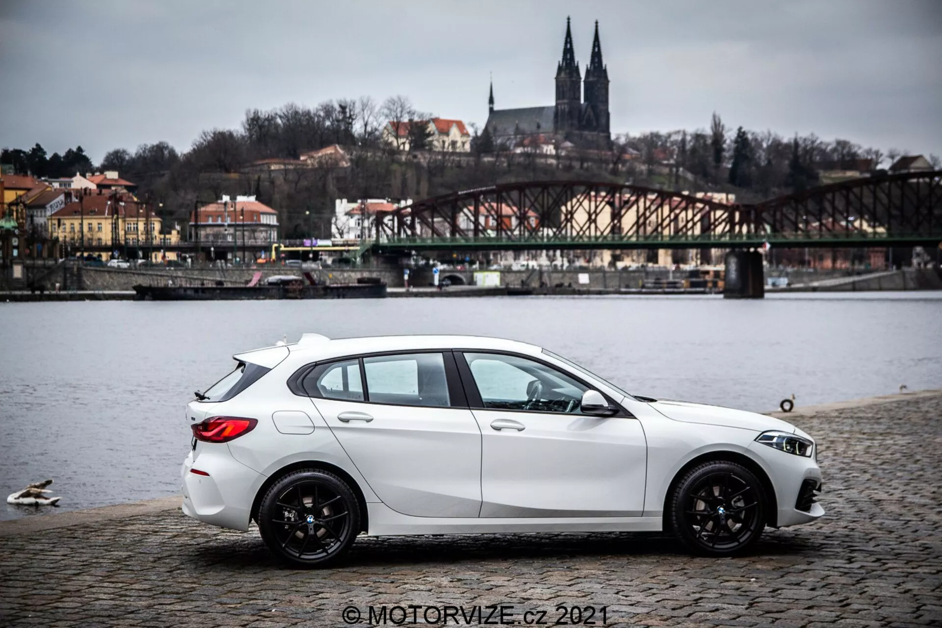 Biely hatchback BMW radu 1 z roku 2019 (BMW-116i) zaparkovaný na dlaždenom povrchu blízko vodného toku s kačkami, zobrazený v bočnom profile s miernym zadným kvadrantom. Auto má dynamické tvarované línie, klesajúcu strechu, čierne viacramenné zliatinové kolesá a viditeľné svetlomety a bočné zrkadlá. Na pozadí je most, budovy na kopci a architektonické prvky naznačujúce európske prostredie. Osvetlenie je rozptýlené za oblačného neba a obraz obsahuje vodotlač © MOTORVIZE.cz 2021.