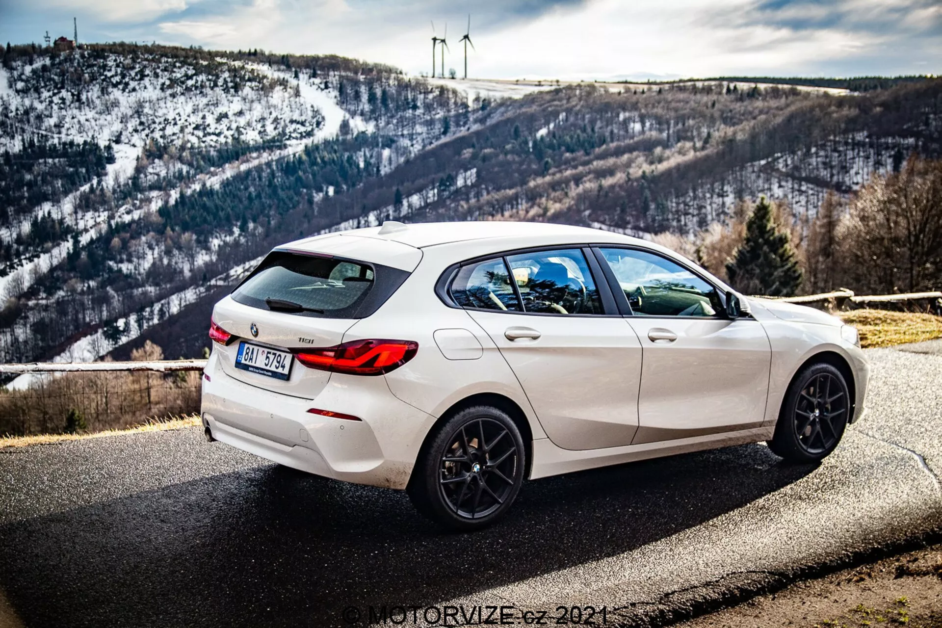 Obrázok ukazuje biely BMW 1 Series (BMW-116i) model 2019 zaparkovaný pri ceste s malebným pozadím zahŕňajúcim kopcovitú, zasneženú krajinu a veterné turbíny na obzore. Fotografia je urobená zo zadného trojštvrťového uhla, zreteľne zobrazujúca pravú stranu vozidla. Pohľad ukazuje elegantné dizajnové línie, zadný spojler, čierne päť-lúčové zliatinové kolesá, dvojité výfuky a tmavé súkromné ​​sklá na zadných oknách. Auto má tiež zadný stierač a integrovanú anténu v tvare plutvy žraloka na streche. Hĺbka poľa drží auto v ostrosti za denného svetla, zvýrazňujúc jeho športovú estetiku, zatiaľ čo pozadie zostáva jemne detailné.