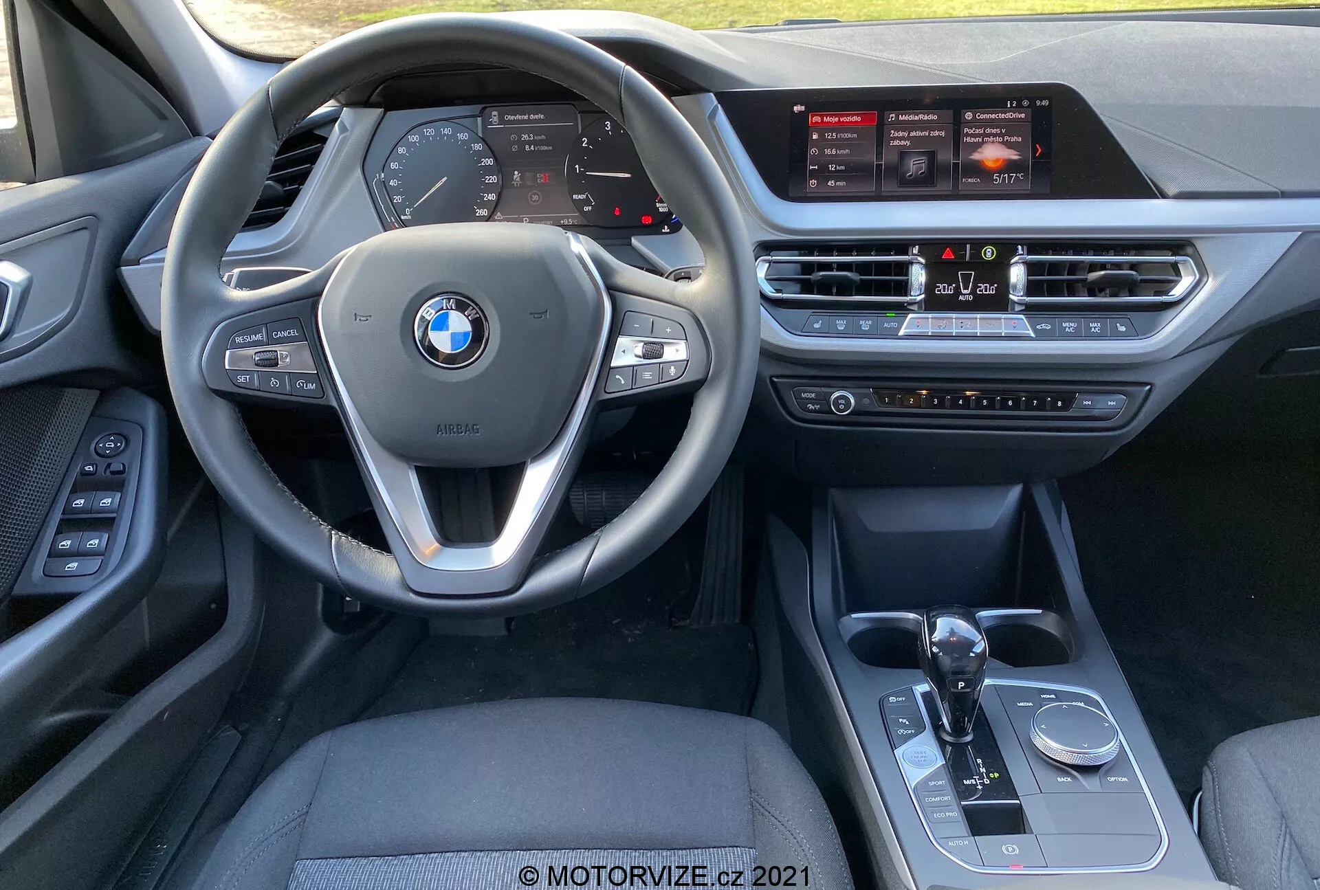 Pohľad do interiéru BMW 1-er reihe (BMW 116i) 2019 z perspektívy sedadla spolujazdca, ktorý ukazuje multifunkčný volant s ovládacími prvkami, digitálny prístrojový panel, veľkú dotykovú obrazovku infotainmentu, systém klimatizácie, páku automatickej prevodovky, prepínač jazdných režimov a ovládací gombík iDrive.