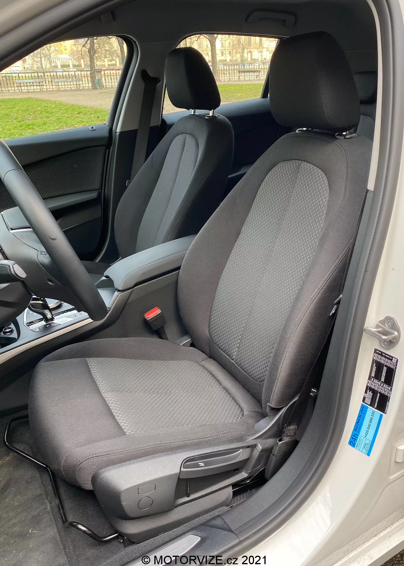 Detailný záber interiéru BMW 1er-Reihe 2019 (BMW-116i) zameriavajúci sa na sedadlo vodiča s moderným dizajnom látky v dvojfarebnom prevedení, prevažne čiernym so svetlejšími šedými vzormi, zo strany s dverami auta široko otvorenými. Sedadlo má opierku hlavy nasmerovanú proti svetlu, čím vytvára siluetový efekt okolo okrajov, a obsahuje páčku na manuálne nastavenie dopredu alebo dozadu, ako aj malé čierne tlačidlo pri okraji interiéru auta. Prirodzené denné svetlo podčiarkuje ostrý zaostrenie na sedadlo, s rozmazaným pozadím zobrazuje volant a prístrojovú dosku.