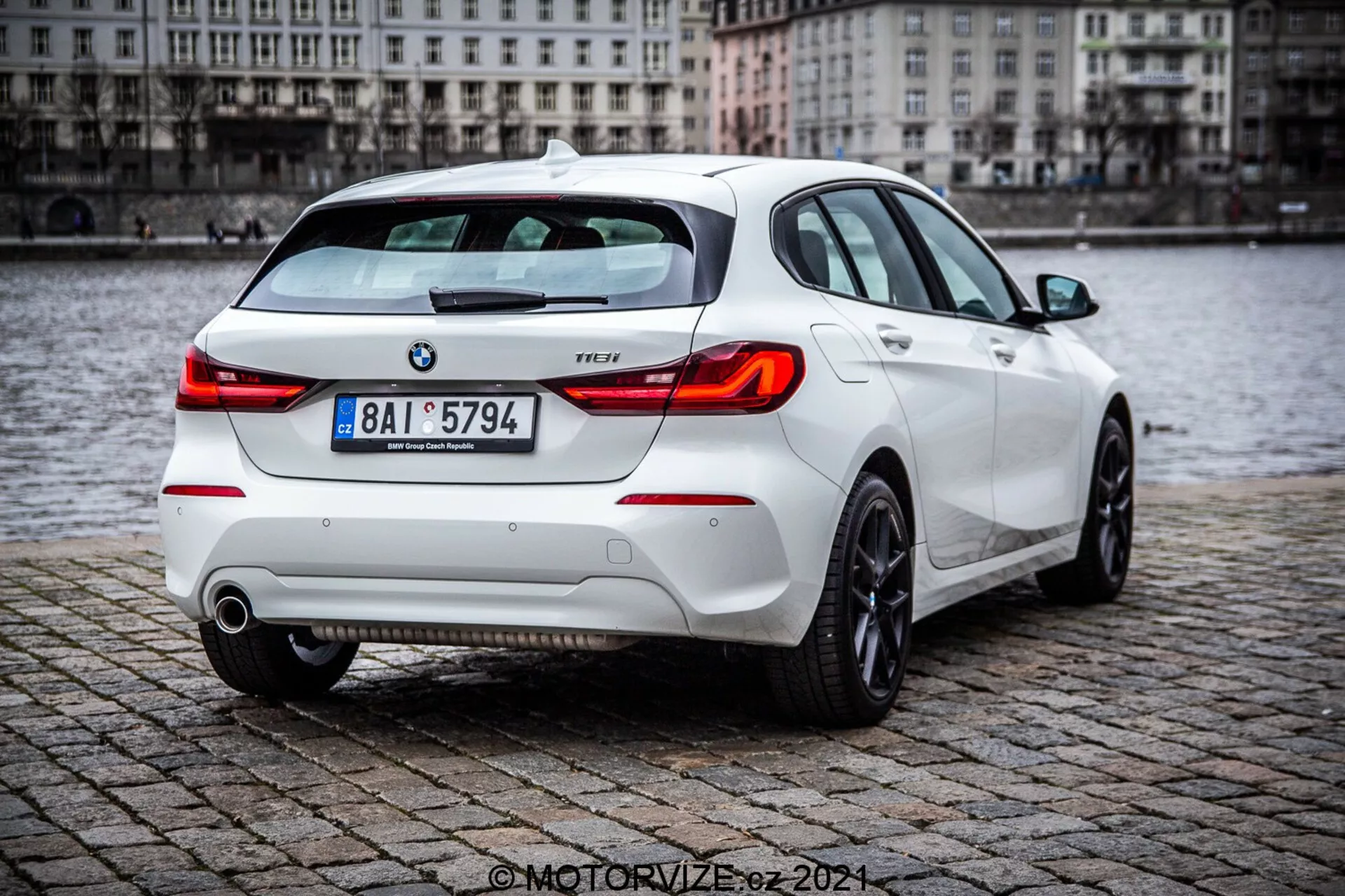 BMW radu 1 z roku 2019 (116i) biely hatchback, zobrazovaný z trojštvrtečného zadného uhla s detailnými prvkami vrátane zadných svetiel, veka kufra, zadného nárazníka, výfukových rúrok a časti zadných kolies, zaparkovaný na mokrej dlažbe blízko vodnej plochy. Obrázok zvýrazňuje športový zadný spoiler, moderné zadné svetelné klastre a viditeľné znaky BMW a úroveň výbavy.