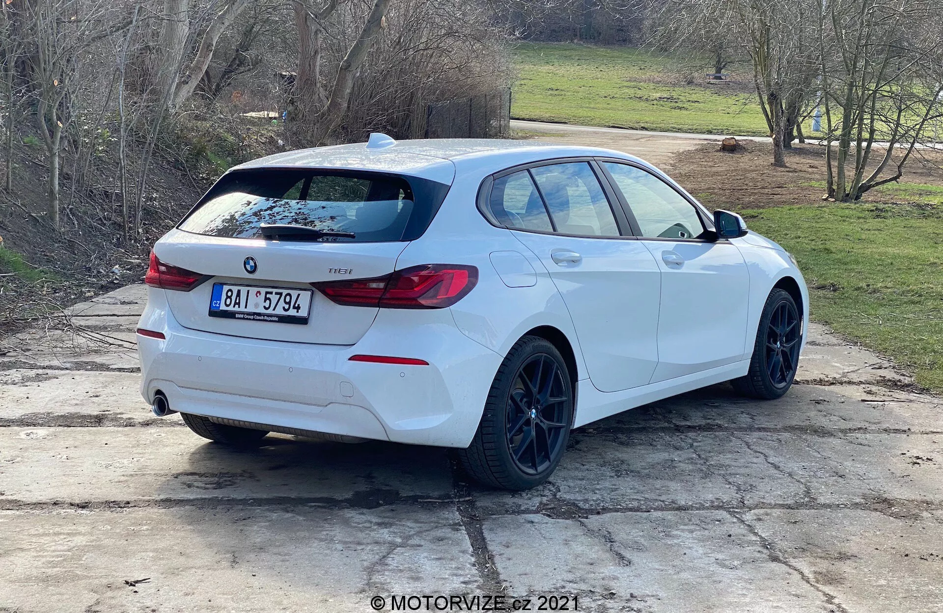 Zadný trojštvrťový pohľad na biely hatchback BMW 1 Series (model BMW 116i z roku 2019) zaparkovaný vonku počas dňa. Profil a zadná časť vozidla sú jasne viditeľné, s emblémom BMW, označením modelu, charakteristickými zadnými svetlami obopínajúcimi zadnú stranu, zadným spojlerom, čiernymi ráfikmi, hatchbackovým karosárskym štýlom, dvojitými výfukovými rúrami, zadným stieračom, jemne tónovanými oknami a spätnými zrkadlami s integrovanými ukazovateľmi smeru. Auto stojí na betónovej ploche s trávnatými škvrnami a vidieckym alebo prímestským prostredím v pozadí.