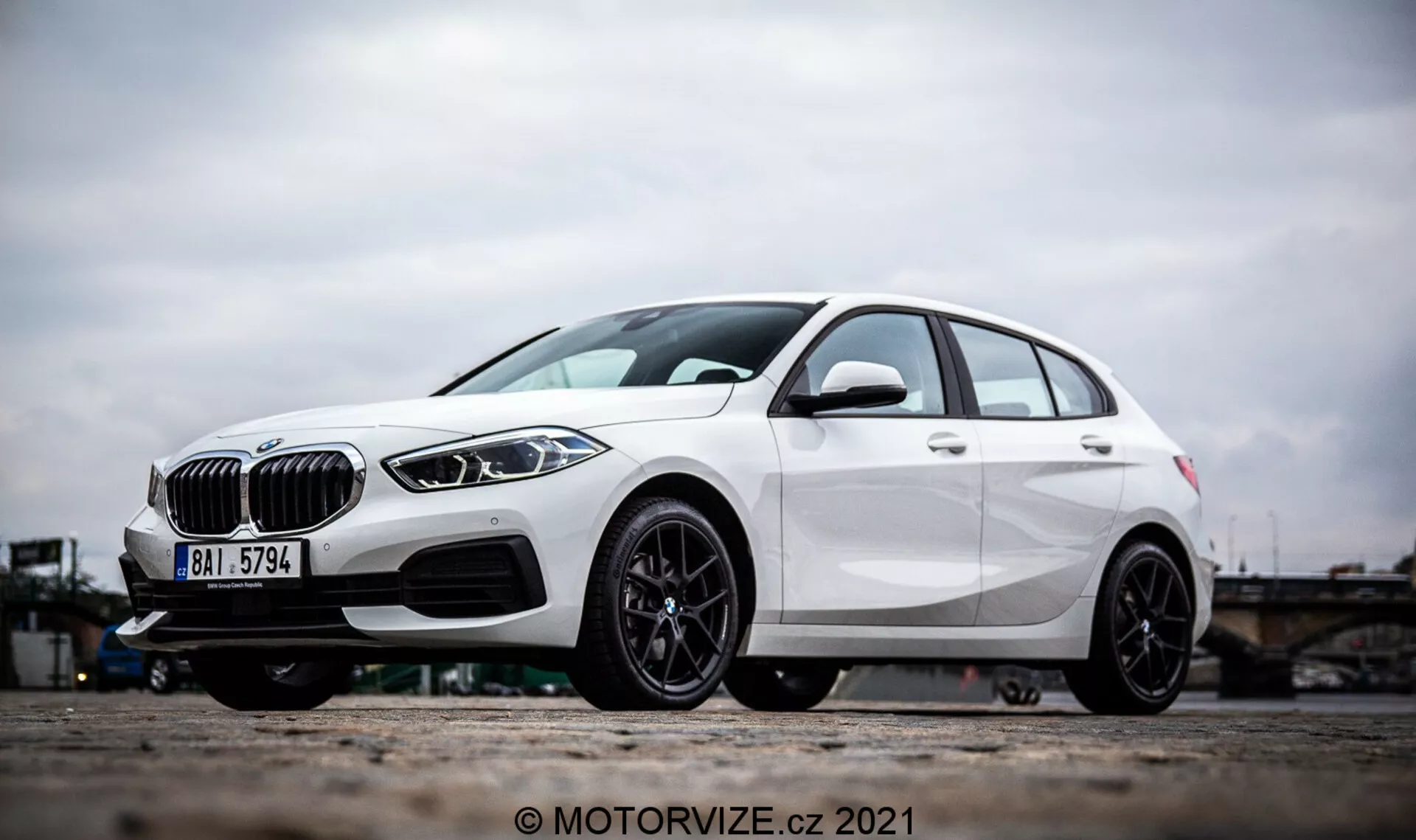 Šikmý bočný pohľad na biele BMW 1er rad 2019 (BMW 116i), ktorý zdôrazňuje výrazné dizajnové prvky, ako je ladvinkovitá mriežka, elegantné LED svetlomety a predný nárazník s otvormi na prísun vzduchu a možnými radarovými senzormi pre asistenčné systémy vodiča. Fotografia sa zameriava na zliatinové kolesá a spodnú časť vozidla, zdôrazňujúc športový čierny viacramenný dizajn a výkonný kotúčovú brzdu. Charakteristické línie a aerodynamické tvarovanie hatchbacku ďalej zdôrazňujú jeho moderný a dynamický exteriér.