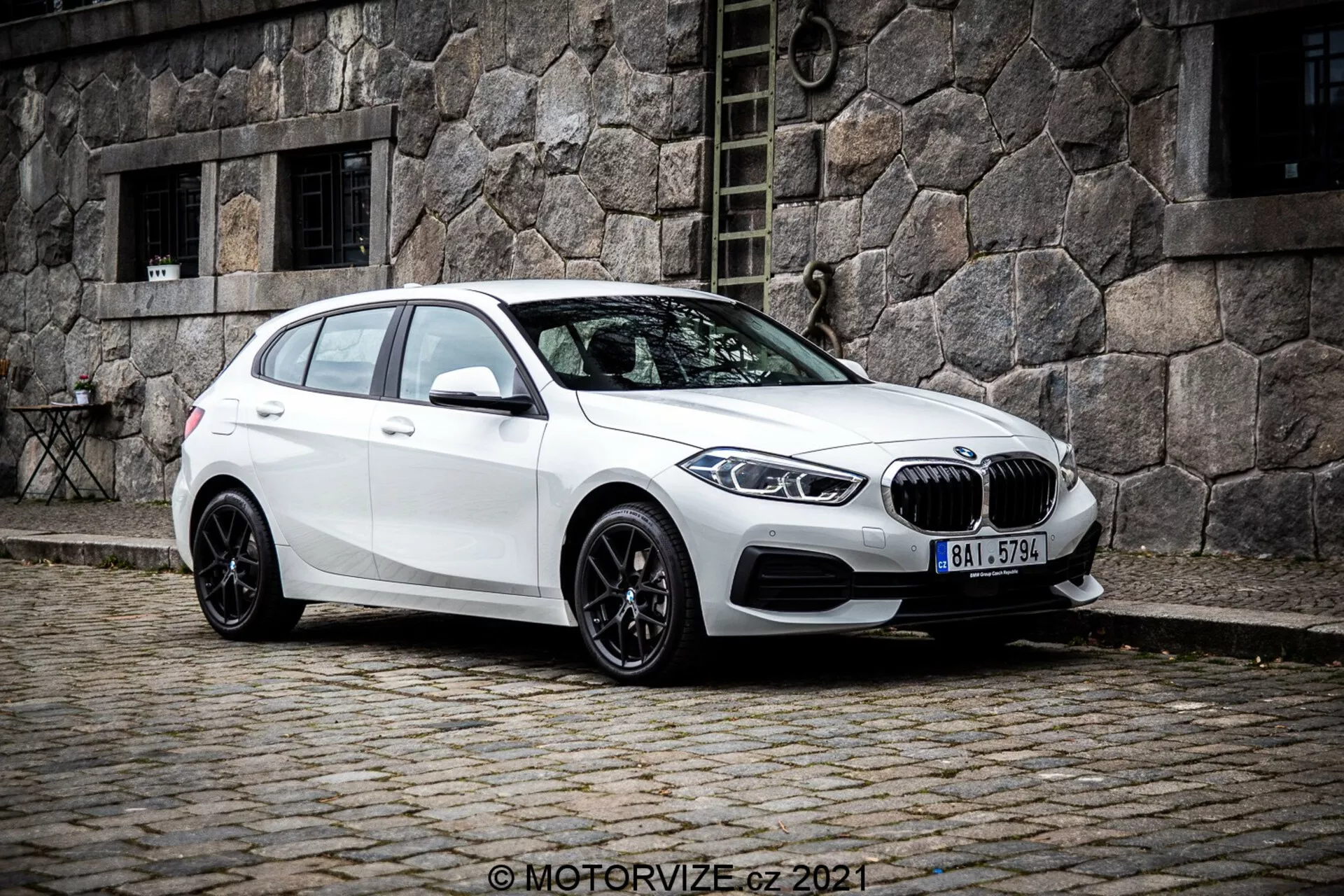 Biely hatchback BMW 1er-Reihe 2019 (BMW 116i) zobrazený z trojštvrťového čelného uhla, ukazujúci ľavú stranu vozidla. Fotografia, urobená z mierne nižšieho uhla, zvýrazňuje postoj a dizajnové línie auta. Je umiestnené na dlažobnej ulici s historickou kamennou budovou v pozadí, vytvárajúc klasickú mestskú scénu. Predná časť s charakteristickou obličkovou mriežkou a BMW emblémom je výrazná, obklopená elegantnými šikmými LED svetlometmi. Auto je vybavené viacramennými čiernymi zliatinovými kolesami, naklonenou strešnou čiarou, bočnými zrkadlami vo farbe karosérie s integrovanými smerovkami a tónovanými oknami pre zadných pasažierov.