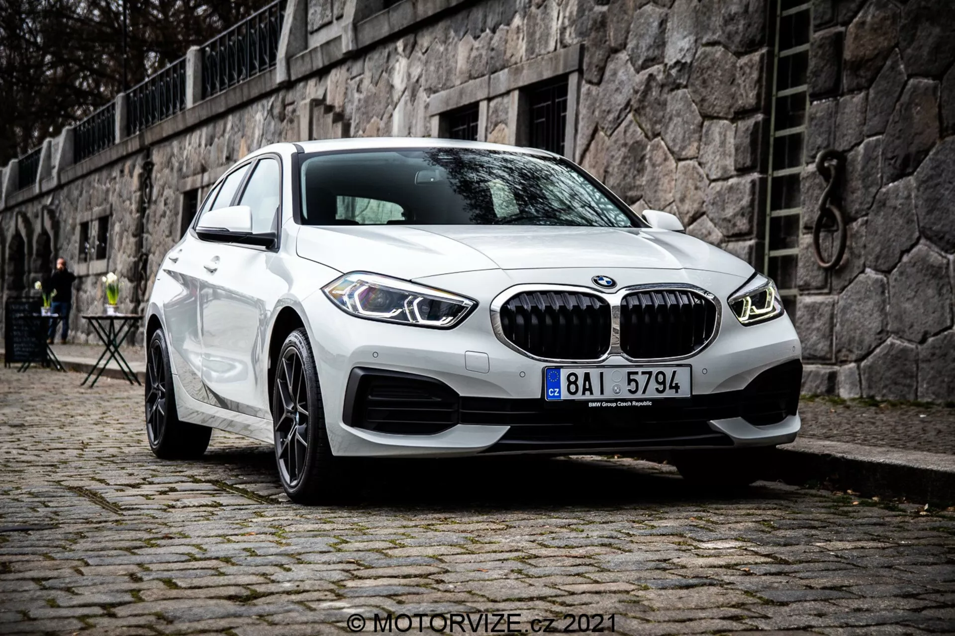 Moderné biele BMW 1. rady (BMW 116i) model 2019 s charakteristickou obličejovou maskou, dvojitými kruhovými LED dennými svetlami a športovým dizajnom predného nárazníka, zaparkované na dláždenej ploche s historickým európskym prostredím so kamenným múrom a vonkajším kaviarenským nábytkom v pozadí.