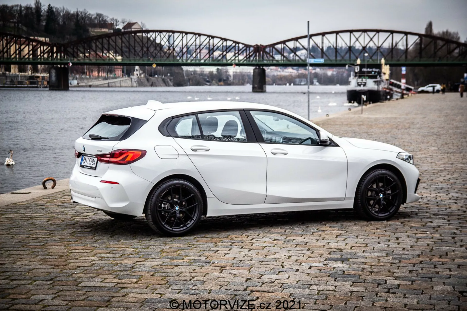 Obrazok bieleho hatchbacku BMW 1er-Reihe 116i z roku 2019 z pohľadu zozadu z trojštvrtinového uhla, zdôrazňujúc jeho elegantný, moderný dizajn so športovým postojom, čiernymi viacramennými zliatinovými kolesami, mierne tónovanými oknami a praktickými funkciami, ako je odmrazovač zadného okna a stierač. Auto zobrazuje ostré línie a kontúry, dvojitý výfukový systém, jemný zadný strešný spojler, zadnú kameru a parkovacie senzory. Je zaparkované na dlažobných kockách v blízkosti vodnej plochy s mestským mostom v pozadí, čo naznačuje kontext vonkajšieho životného štýlu.