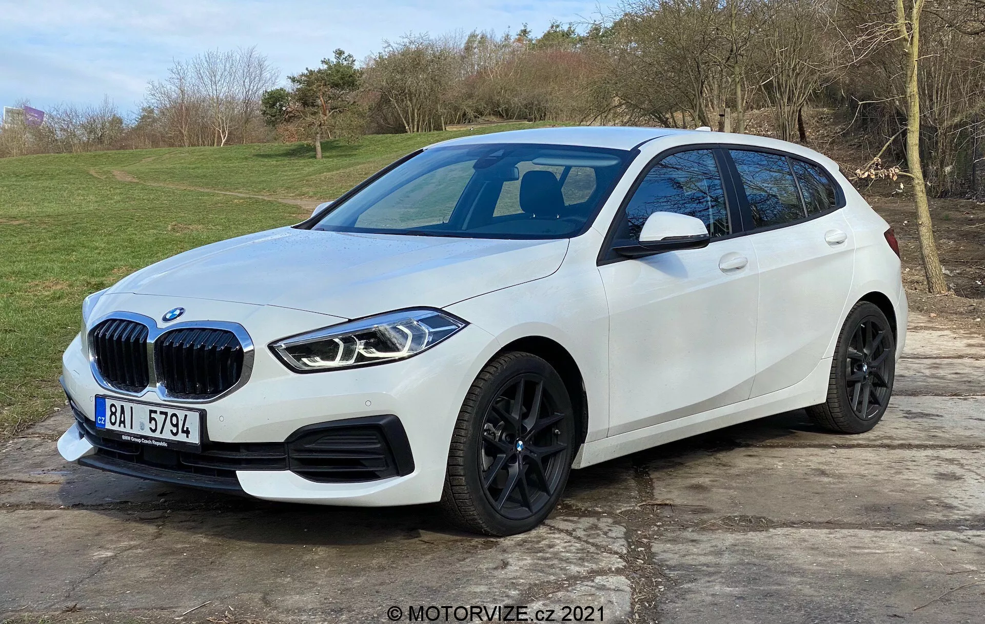 Obrázok zobrazuje moderný biely hatchback BMW rad 1 (BMW 116i) umiestnený v exteriéri s prírodným trávnatým pozadím. Zobrazený z predného trojštvrťového pohľadu, uprednostňujúceho stranu vodiča, ukazuje charakteristickú mriežku v tvare obličiek vozidla, elegantné hranaté LED svetlomety, čierne zliatinové kolesá, dobre definovaný bočný záhyb a integrované smerové svetlá na bočných zrkadlách. Predný nárazník obsahuje parkovacie senzory, zatiaľ čo panoramatická sklenená strecha a prípadná predná kamera sú tušené. Auto je zaparkované na dláždenej ploche a zaberá väčšinu rámu, čo naznačuje moderný automobilový dizajn a pokročilé technologické prvky.