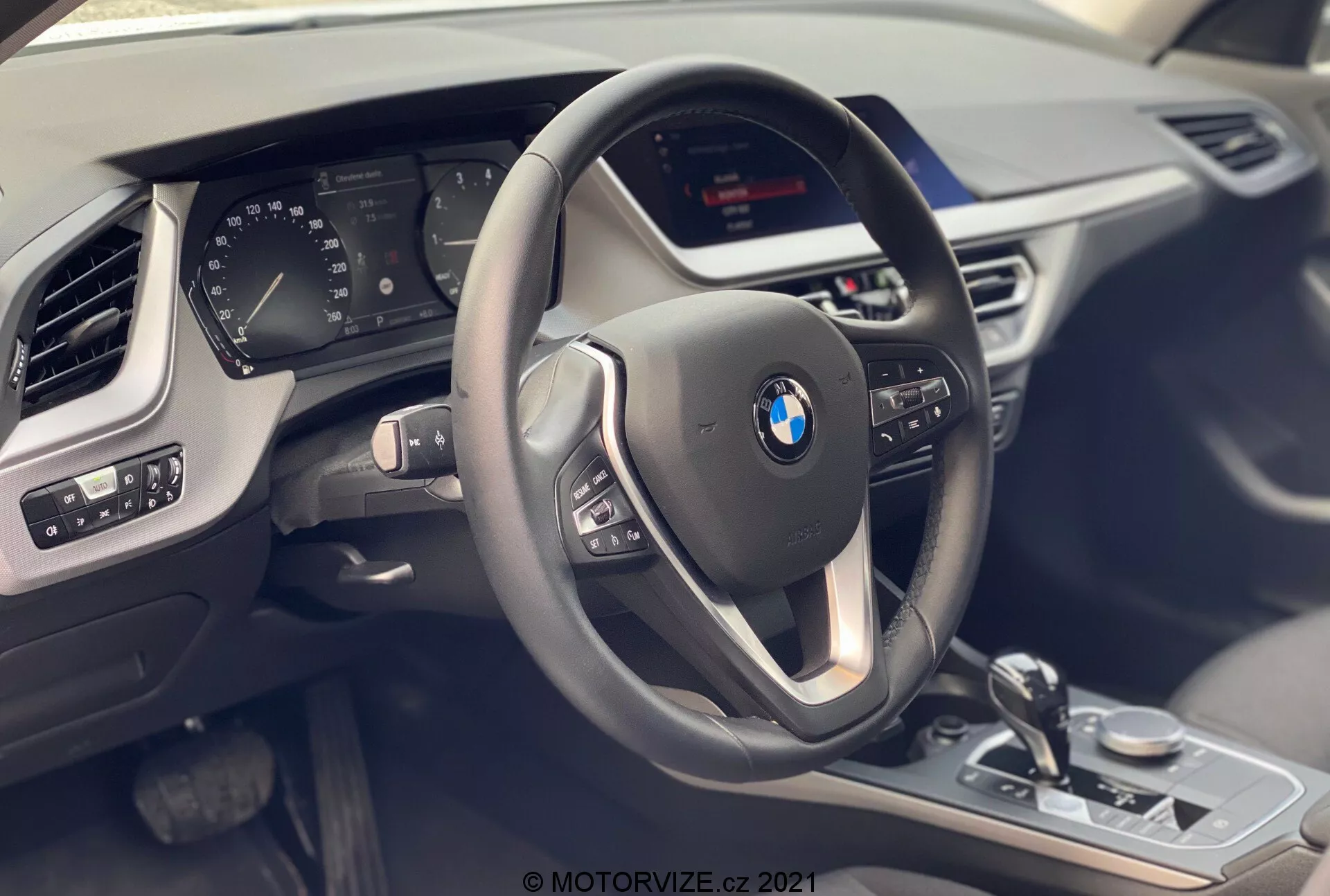 Pohľad na interiér BMW 1 Series 2019 (BMW 116i) zo sedadla spolujazdca, zdôrazňujúci vodičský kokpit s moderným trojramenným volantom s multifunkčnými tlačidlami a koženým poťahom s kontrastným prešívaním. Za volantom je čiastočne viditeľný digitálny prístrojový panel zobrazujúci rôzne parametre vozidla. Stredová konzola obsahuje neviditeľnú obrazovku infotainmentu a viditeľné ovládacie prvky, ako je voliaca páka prevodovky, ovládacia gombík iDrive, prepínač jazdných režimov a ovládanie parkovacích senzorov, všetko s kovovými akcentmi a lesklou čiernou úpravou. Ovládací panel dverí vodiča ukazuje nastavenia zrkadiel a elektrické ovládače okien. Viditeľná je aj časť ventilačného otvoru na strane spolujazdca a textúra prístrojovej dosky, ktorá zdôrazňuje luxus, technológiu a moderný dizajn.