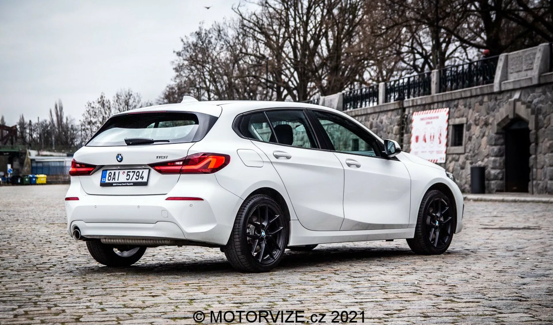 Vysokokvalitná fotografia bieleho hatchbacku BMW 1 Series 2019 128ti, fotografovaného zo zadného trojštvrťového uhla, ktorý zvýrazňuje pravú stranu. Auto je zaparkované na dlaždenom povrchu s kamenným múrom v pozadí, čo zdôrazňuje jeho športový dizajn so zadným strešným spojlerom, dvojitou výfukovou sústavou, zadným difúzorom, LED zadnými svetlami, zatmavenými oknami a tmavými zliatinovými kolesami. Rozptýlené osvetlenie, absencia ostrých tieňov a jasné detaily naznačujú zamračené prostredie, poskytnúc autentickú reprezentáciu farby a funkcií vozidla. Obrázok nesie vodoznak 'MotorVize.cz 2021'.