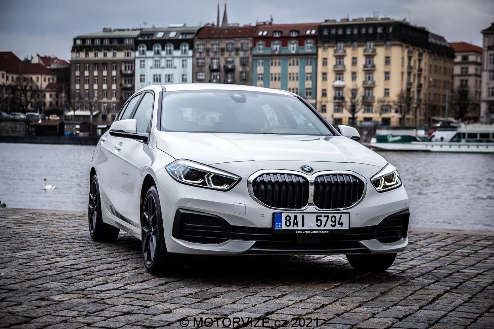 Obrázok zobrazujúci BMW 1er-Reihe 2019 (BMW 116i) z prednej trojštvrťovej perspektívy, zvýrazňujúci obrysy a dizajnové prvky moderného luxusného sedanu. Auto je umiestnené vonku na dláždenom násype pri vodnej ploche, pod zamračenou oblohou. Výrazné prvky zahŕňajú mriežku s vertikálnymi lamelami, elegantné LED svetlomety, integrované hmlové svetlá, zliatinové kolesá a aerodynamický profil.