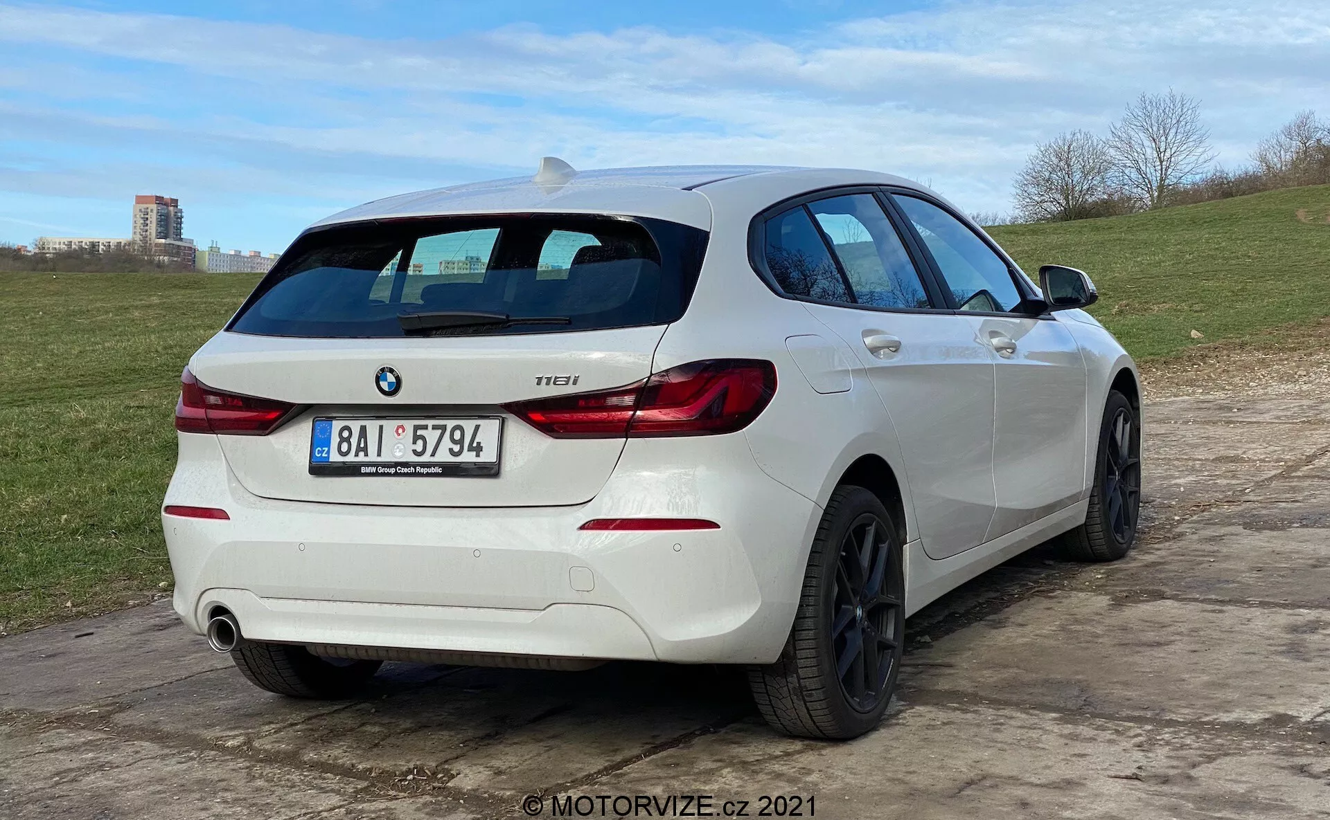 Pohľad z troch štvrtín zozadu na biely hatchback BMW 1 Series 118i, zaparkovaný na nerovnom povrchu. Auto má charakteristické zadné svetlá tvaru „L“, odznak BMW a modelový odznak s označením „118i“. Má dvojitý výfukový systém s jednou viditeľnou rúrkou na pravej strane zadného nárazníka. Vozidlo je vybavené čiernymi zliatinovými kolesami s viac lúčmi a zadným stieračom. Strešný spojler dodáva športový vzhľad a bočný profil odhaľuje ľahko tónované okná, pohlcovacia karosérie a kľučky a zrkadlá zodpovedajúce farebnej schéme auta. Nízky uhol kamery zdôrazňuje postoj auta a dizajn kolies.