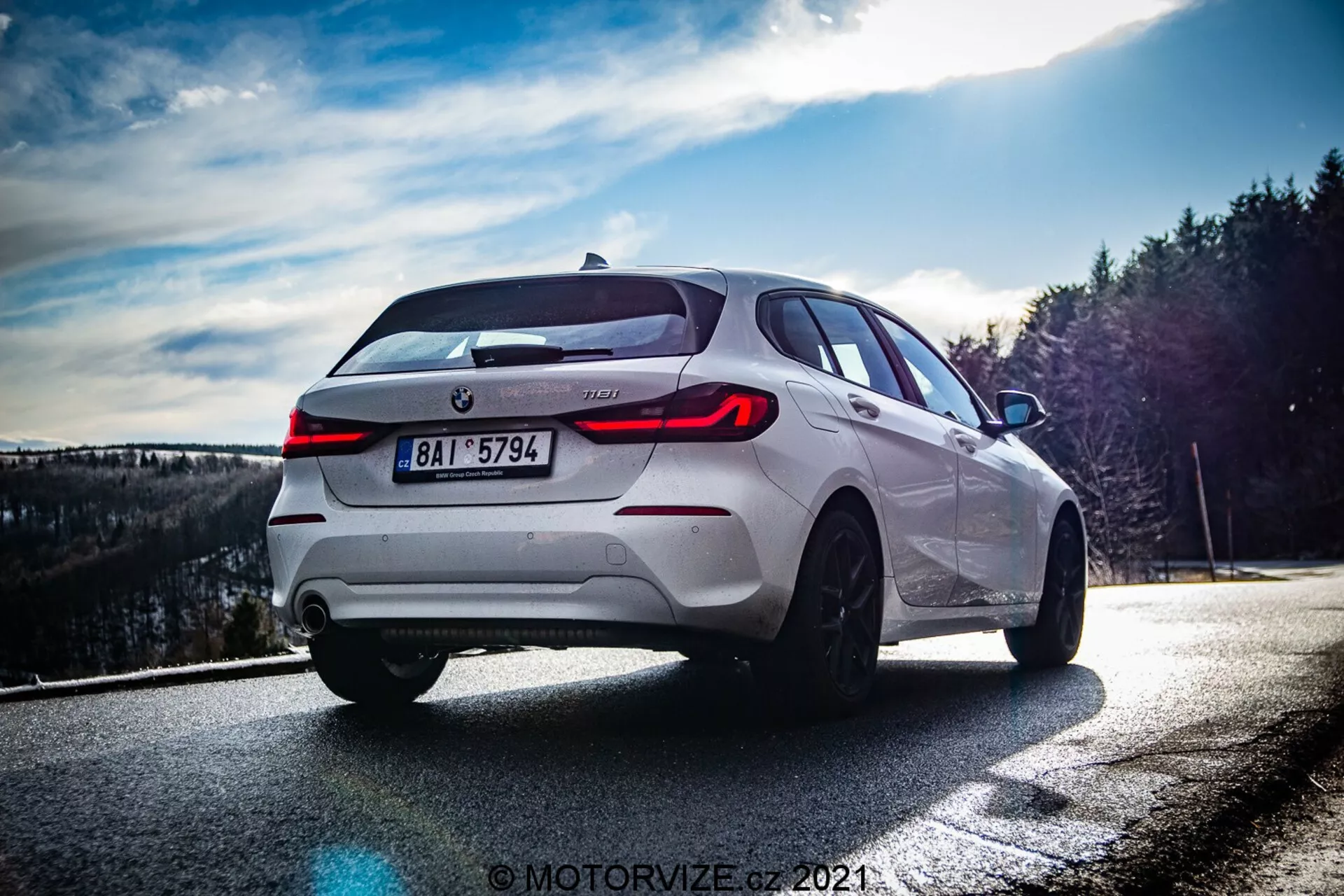 Obrázok zobrazuje biely BMW radu 1 2019 (BMW-116i) zachytený z nízkeho a čiastočne zadného trojštvrťového uhla, zdôrazňujúci zadnú pravú stranu vozidla. Denné svetlo, pravdepodobne neskoré popoludnie, vytvára dynamické kontrasty, ktoré zvýrazňujú dizajnové línie. Výrazné prvky zahŕňajú výrazné LED zadné svetlá, centrálny znak výrobcu a označenie modelu, dvojitý výfukový systém, viaclúčové zliatinové kolesá, pneumatiky s nízkym profilom, viditeľné kotúčové brzdy a športovú siluetu hatchbacku s nakloneným zadným sklom a strešným spojlerom. Kompozícia a svetelné efekty fotografie dodávajú dramatický nádych, rozmazané pozadie sústreďuje pozornosť výlučne na vozidlo.