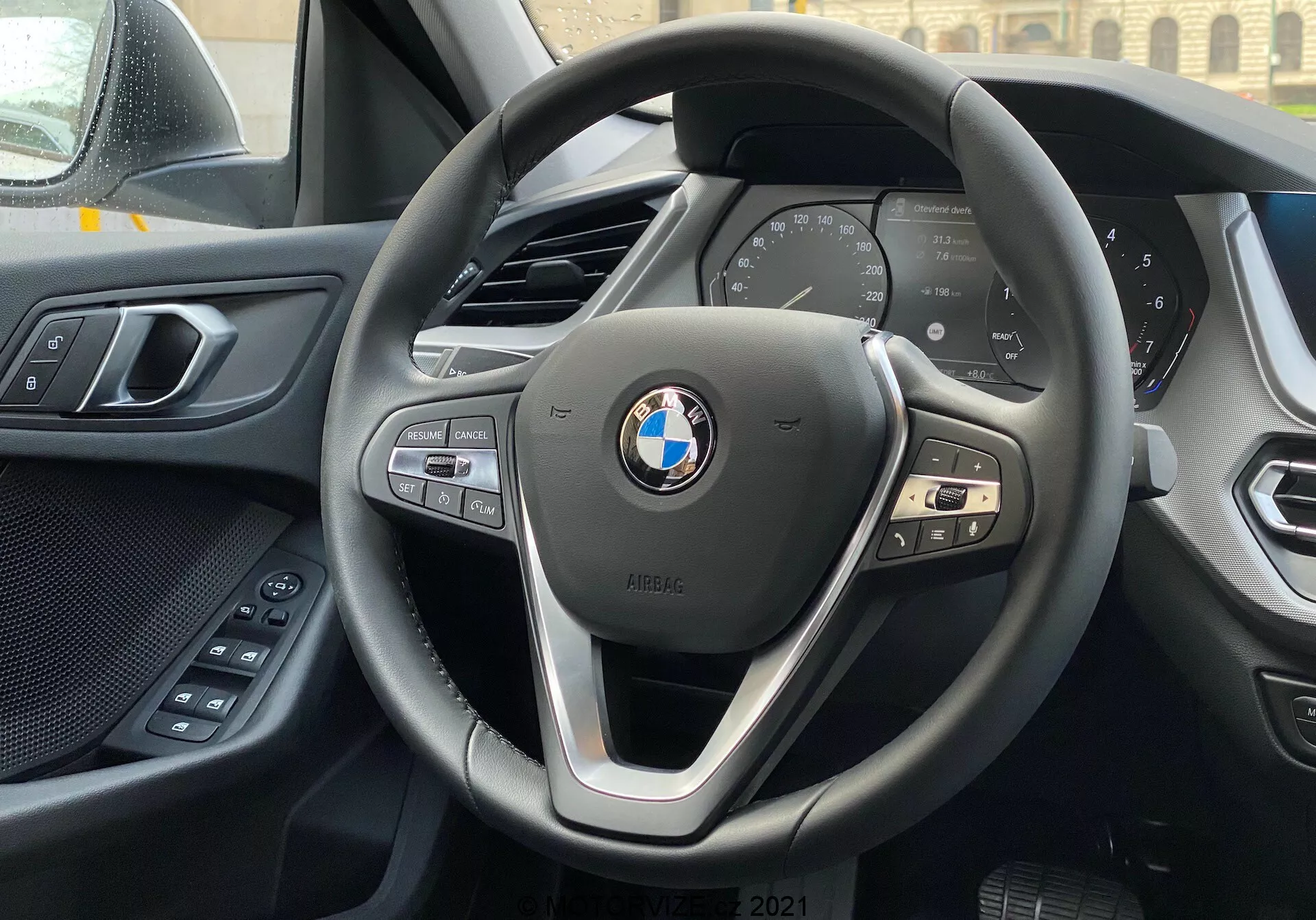 Pohľad na interiér vozidla BMW 1. série 2019 (BMW 116i) zo sedadla vodiča so zameraním na moderný trojramenný volant BMW s ovládacími tlačidlami pre multimédiá, telefón a tempomat a digitálny prístrojový panel zobrazujúci základné údaje o vozidle, ako sú rýchlosť a otáčky. V pozadí je vidieť hornú hranu stredovej konzoly a naľavo časť dverí vodiča s elektrickými ovládacími prvkami pre pohodlie. Prístrojová doska a panel dverí ukazujú kombináciu mäkkých materiálov a kvalitného spracovania. Emblém BMW a odznak iPERFORMANCE sú na volante výrazne zobrazené, čo naznačuje efektívnosť alebo hybridnú technológiu.