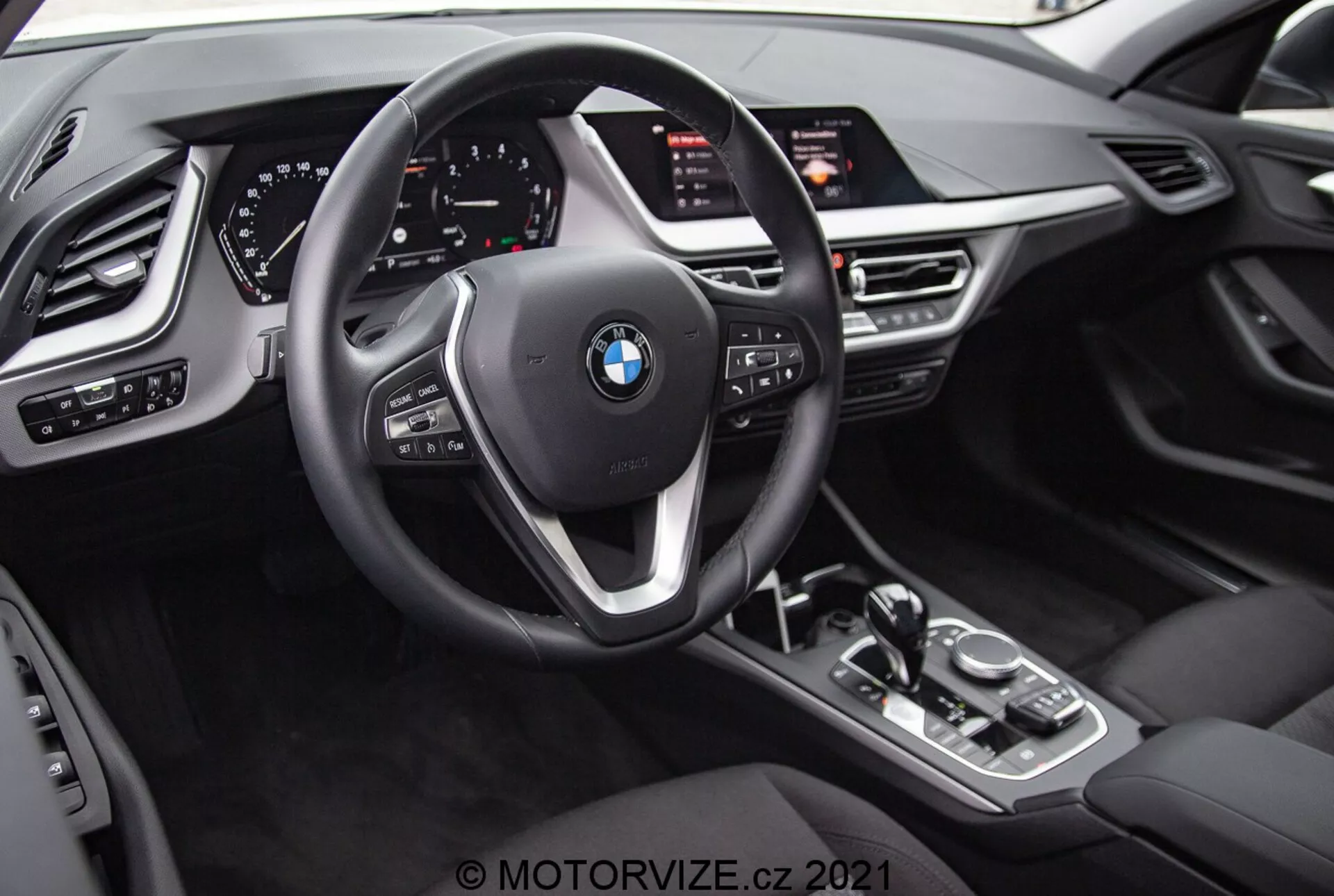Pohľad do interiéru BMW 1er-reihe 2019 (BMW 116i) z perspektívy vodiča. Obrázok zvýrazňuje emblém BMW na trojramennom volante s multifunkčnými tlačidlami a radiacimi pádlami za volantom. Za volantom je plne digitálny prístrojový panel so svietiacimi ciferníkmi a rôznymi informačnými displejmi. Stredová konzola obsahuje radiacu páku automatickej prevodovky, ovládací gombík iDrive, tlačidlá na jazdné režimy a veľkú infotainment obrazovku, ktorá je mierne naklonená smerom k vodičovi. Dizajn interiéru zahŕňa kožený vzhľad, kovové akcenty a ďalšie ovládače pre svetlá, okná a nastavenie zrkadiel s pamäťovou funkciou pre vodičove sedadlo.