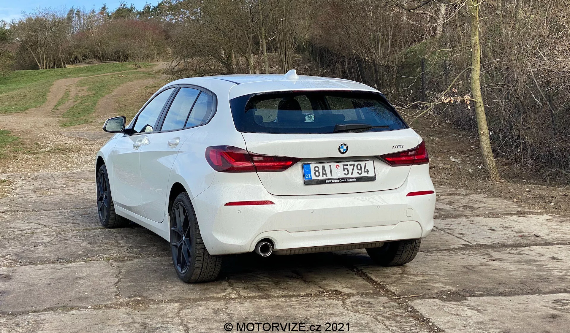 Obrázok zobrazuje biely hatchback BMW 1er-reihe 2019 (BMW 116i) z uhla zadnej tretiny vozidla. Zreteľne sú vidieť horizontálne orientované zadné svetlá, dvojitý výfukový systém a parkovacie senzory integrované do zadného nárazníka. Na zadných dverách je viditeľné logo BMW a označenie modelu. Auto je zaparkované na štrkovom povrchu v prírodnom, vidieckom prostredí. Obrázok naznačuje automobilové témy ako dizajn vozidiel, súčasné modely áut, obsah zameraný na BMW, výfukové systémy a technológie parkovacích asistentov. Vodoznak MOTORVIZE.cz 2021 značí pôvod v automobilovej recenzii alebo publikácii.