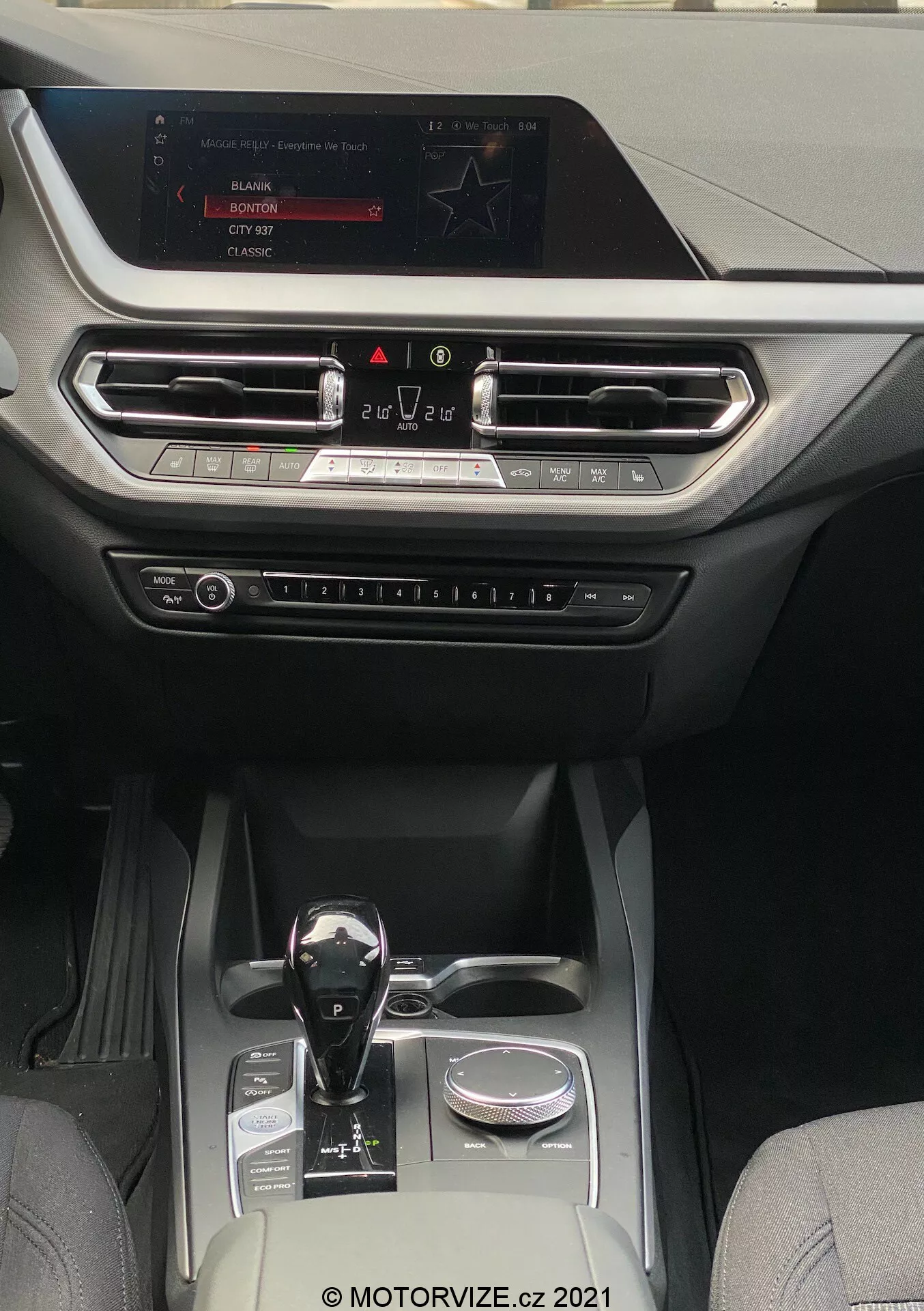 Pohľad na interiér vozidla BMW 1er-reihe 2019 (BMW-116i), ukazujúci modernú a luxusnú kabínu zameranú na stredovú konzolu a oblasť prístrojovej dosky. Perspektíva je z priestoru zadných sedadiel smerom dopredu k infotainment obrazovke umiestnenej hore na prístrojovej doske. Displej zobrazuje možnosti rádia, médií a nastavení vozidla. Pod obrazovkou sú vodorovne zarovnané vetracie otvory s kovovými akcentmi a dekoratívnym prvkom pokrývajúcim celú šírku prístrojovej dosky. Pod ventilačnými otvormi sú viditeľné fyzické tlačidlá a gombíky na ovládanie klimatizácie a iných nastavení. Automatická radiaca páka má lesklý čierny povrch s kovovými akcentmi, umiestnená vedľa otočného ovládača. Stredová konzola obsahuje dva držiaky na poháre, malý úložný priestor a strieborný dekoratívny prvok, čo naznačuje dobre vybavenú kabínu kombinujúcu kožu a vysoko kvalitné syntetické látky, vhodnú pre stredné až vyššie kategórie vozidiel zamerané na technológiu a pohodlie.