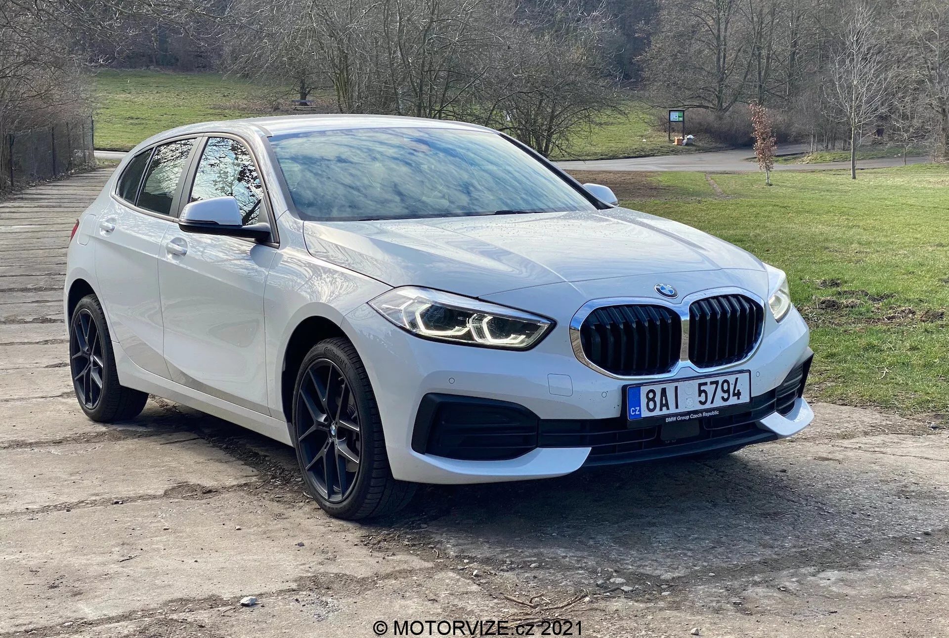 Moderné vozidlo BMW 116i zo série 1 z roku 2019 fotografované z trojštvrťového predného pohľadu, zobrazujúce prednú mriežku s emblémom, elegantné LED svetlomety, hmlové svetlá, predný nárazník a aerodynamické línie kapoty vedúce k čelnému sklu. Koleso a blatník na strane vodiča spolu so zrkadlom s integrovanými smerovkami zdôrazňujú vyspelý dizajn a technológiu parkovacích senzorov vozidla. Zachytené vonku na pozadí stromov a trávy, vozidlo má zliatinové kolesá a charakteristickú mriežku v tvare obličiek, čo naznačuje luxus a výkon.