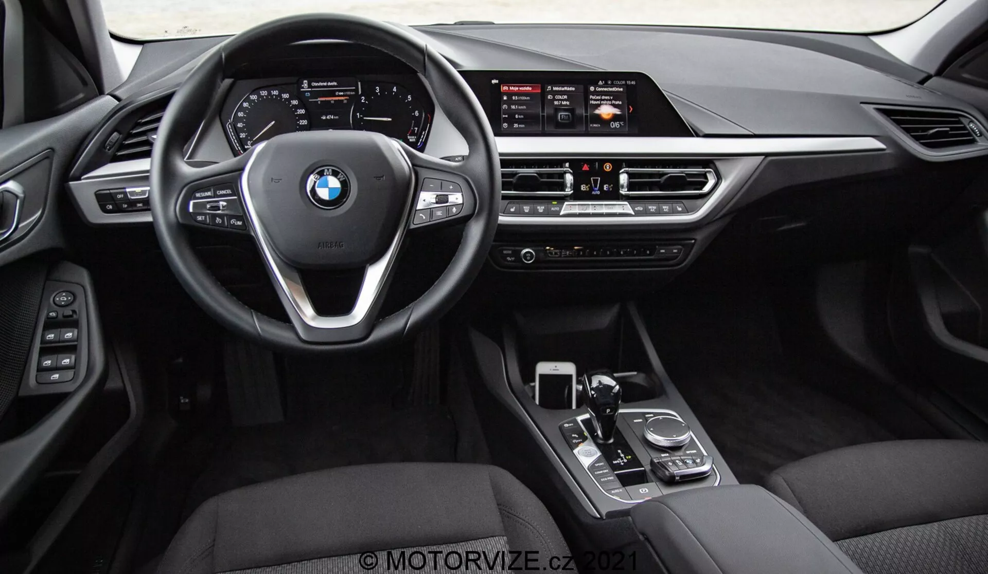 Pohľad na interiér BMW radu 1 2019 (BMW 116i) z pohľadu zadných sedadiel, zobrazujúci celú prednú palubnú dosku, volant s logom BMW a ovládacími tlačidlami a stredovú konzolu. Viditeľné prvky zahŕňajú široký informačný displej, manuálne ovládania klimatizácie, vetracie otvory, radičku prevodovky, otočný volič a touchpad. Interiér sa vyznačuje čiernymi koženými alebo koženkovými prvkami s kovovými a lesklými ozdobnými prvkami, zdôrazňujúcimi luxus a pokročilé technológie pre komfort vodiča.