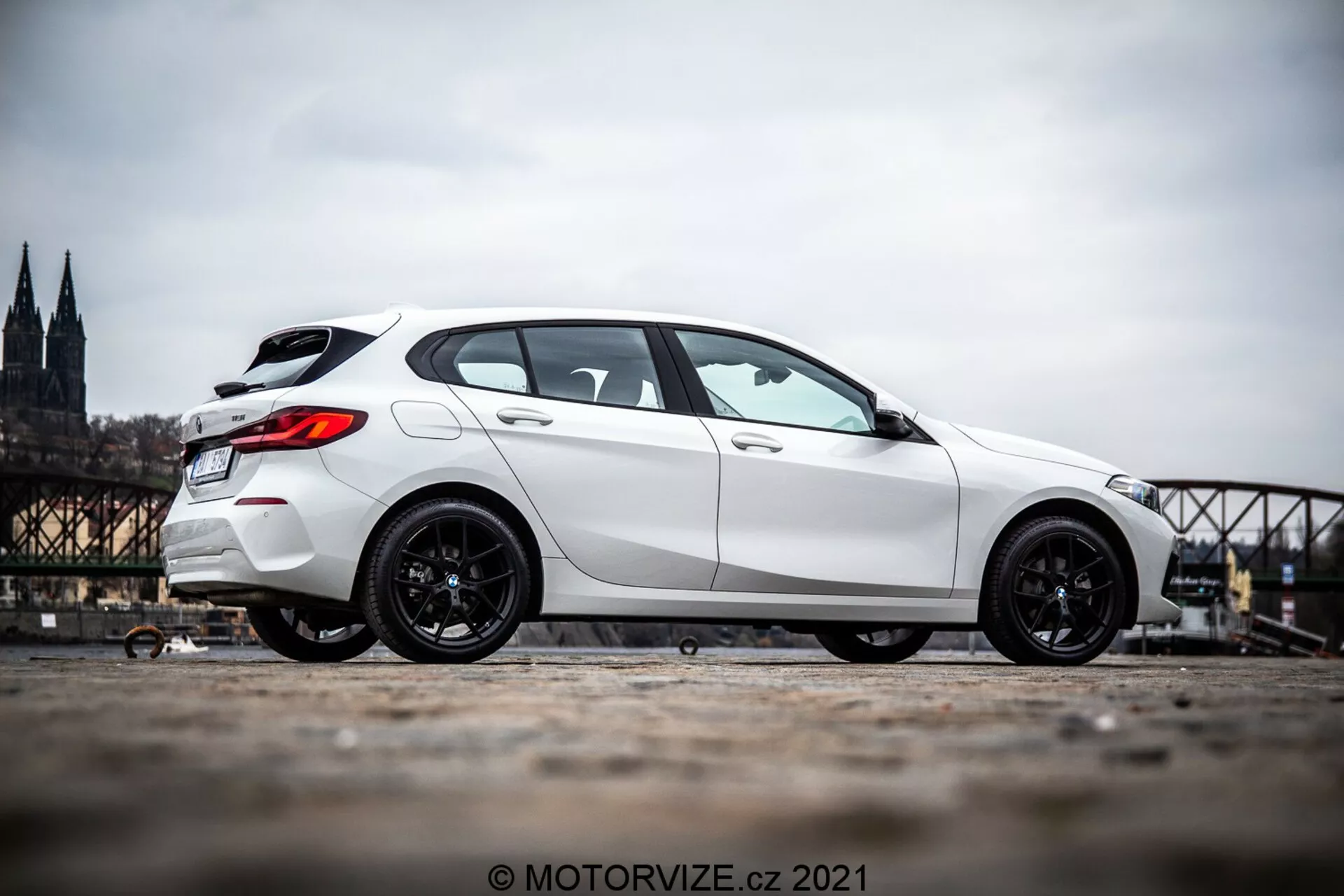 Biely hatchback BMW 1. série (BMW 116i) z roku 2019 fotografovaný z bočného profilu, zobrazujúci bok vozidla od predného kolesa až po zadnú časť. Auto je umiestnené na ľavej strane obrázka s rozmazaným historickým mestským pozadím, ktoré zahŕňa katedrálu a most. Obrázok zvýrazňuje športové tmavo zafarbené ráfiky auta a jeho dominantnú polohu vďaka nižšej úrovni zeme a miernemu nakloneniu kamery nahor. Fotografia je urobená vonku počas dňa a odhaľuje detaily estetického vzhľadu auta v prirodzenom dennom svetle.