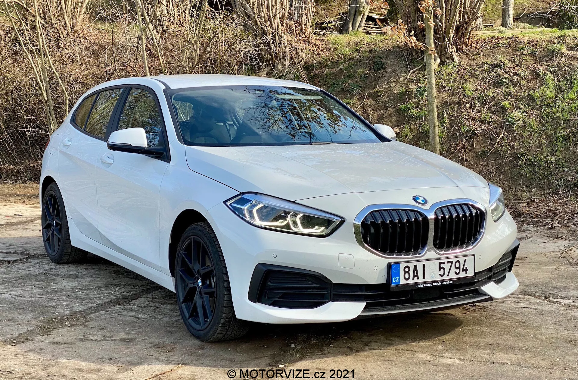 BMW 116i rady 1 z roku 2019 v bielej farbe, zachytený z predného trojštvrťového uhla, ktorý ukazuje jeho výraznú obličkovitú prednú mriežku, uhlové LED svetlá, elegantné čierne kolesá s nízkoprofilovými pneumatikami a aerodynamické línie karosérie. Umiestnený vonku na rovnú plochu s prírodnou vegetáciou v pozadí, záber zvýrazňuje moderný dizajn a športové prvky auta vrátane parkovacích senzorov a bočných zrkadiel s integrovanými ukazovateľmi smeru.