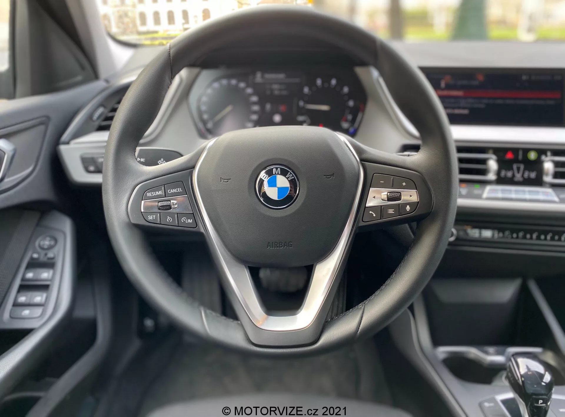 Pohľad z miesta vodiča na BMW 1er-reihe 2019 (BMW-116i), sústredený na moderný trojramenný volant s kovovým akcentom na spodnom rameni a výrazným logom BMW v strede. Na ľavej strane volantu sú ovládacie prvky médií a komunikácie, zatiaľ čo na pravej strane sú tlačidlá tempomatu a viditeľná radiaca páka. Na pozadí je čiastočne viditeľný prístrojový panel so štýlom analógových ukazovateľov a infotainment obrazovkou na stredovej konzole, ktorá naznačuje pokročilé funkcie ako je navigácia GPS a klimatizácia.