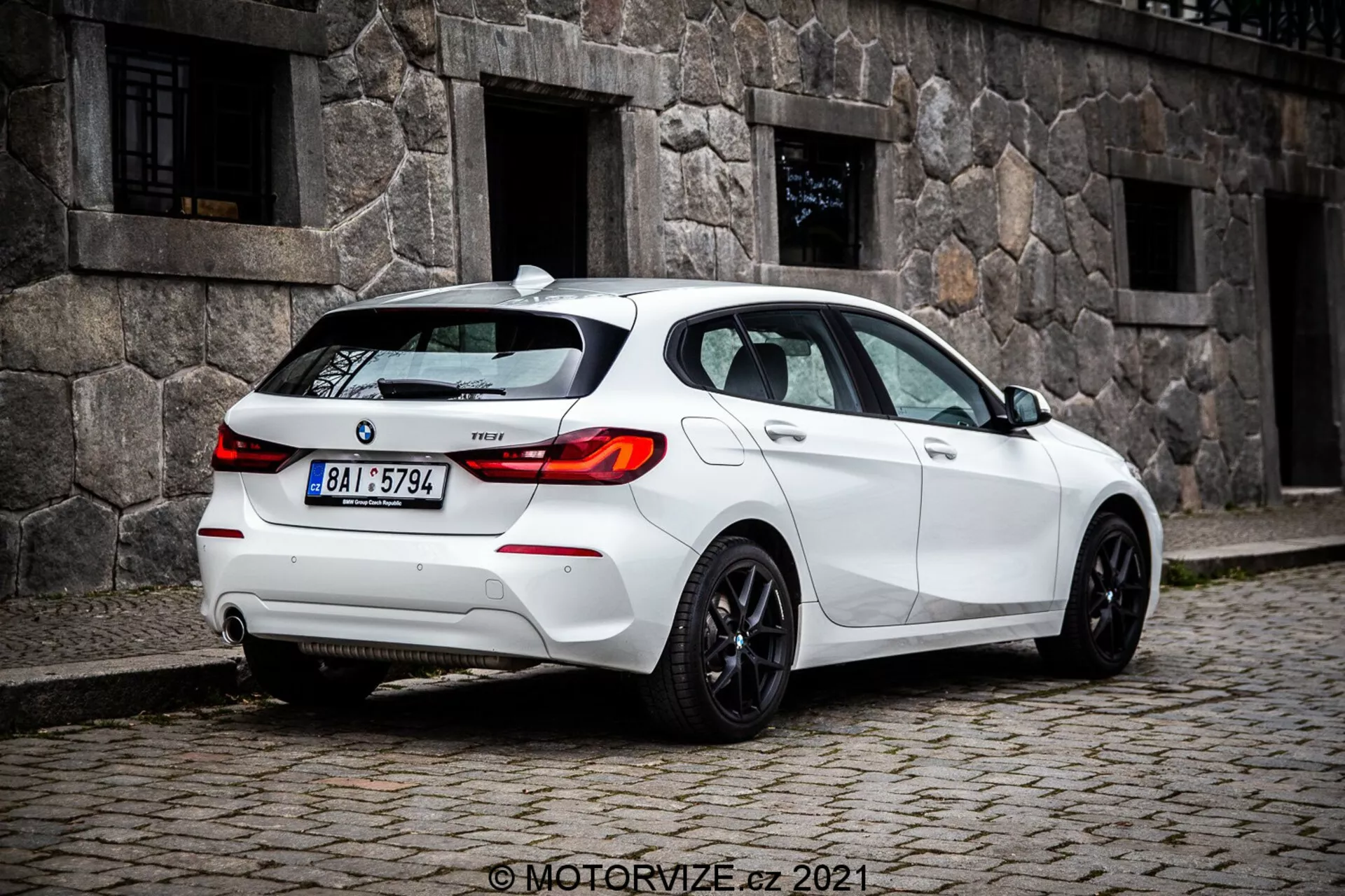 Stredná vzdialenosť a pohľad z úrovne zeme na biely hatchback BMW 1er-rada 116i 2019, zachytený z troch štvrtín zadného pohľadu, výrazne zobrazujúc zadné svetlá s LED dizajnom, zadné sklo a športové disky. Viditeľné prvky zahŕňajú senzory na zadnom nárazníku, pravdepodobne pre parkovacie asistenciu, malú anténu na streche, pravdepodobne pre GPS alebo satelitné rádio, odznak modelu na kufri, dvojitý výfukový systém, ktorý naznačuje výkonnosť, a tónované okná pre súkromie alebo komfort.