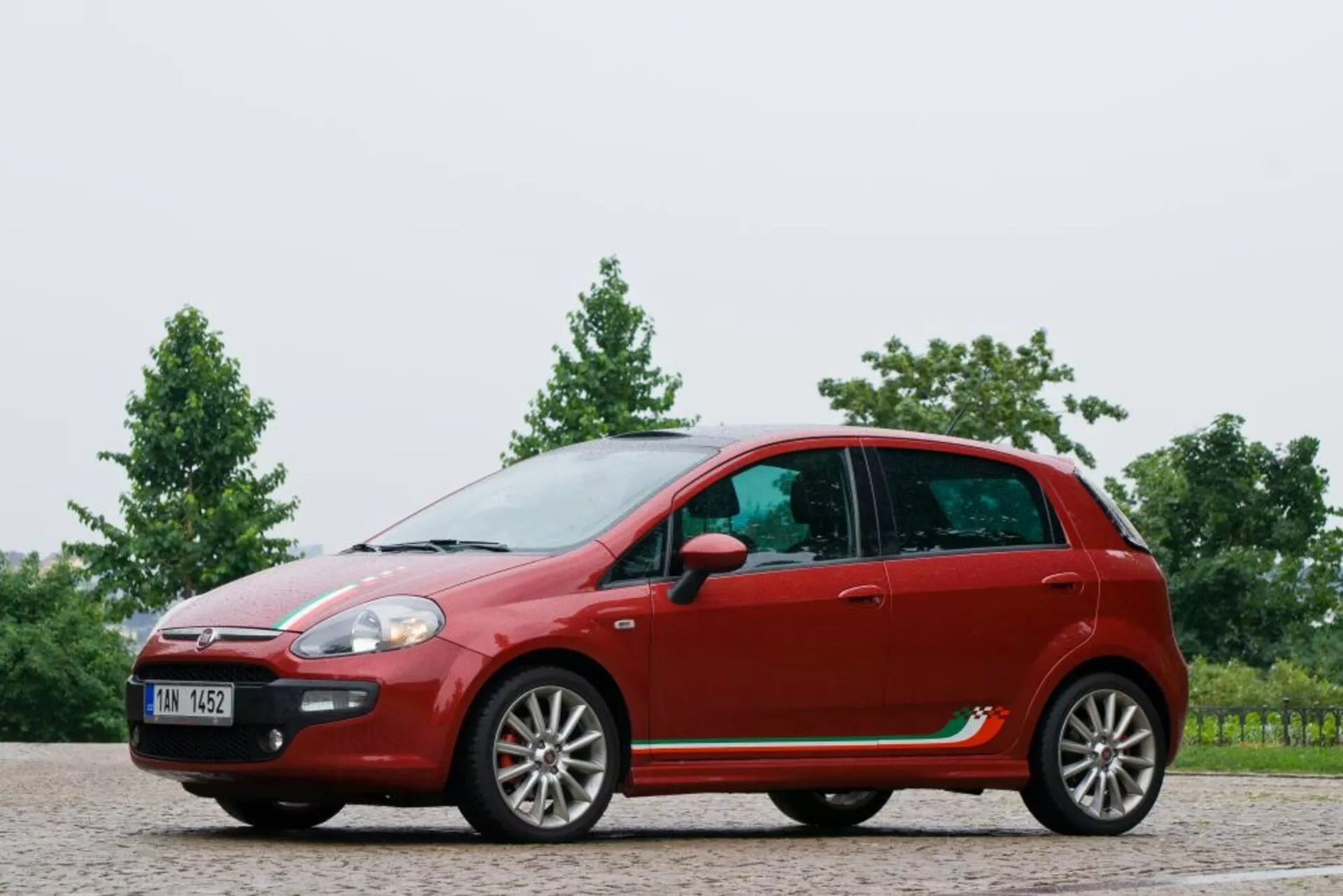 Plný bočný profil červeného hatchbacku Fiat Punto z roku 2011, zobrazujúci jeho zakrivenú strešnú líniu, päťdverovú konfiguráciu s diskami z ľahkej zliatiny, bočné zrkadlá na dverách, výrazné línie charakteru a plastové lišty na štrkovom povrchu so zeleným pozadím.