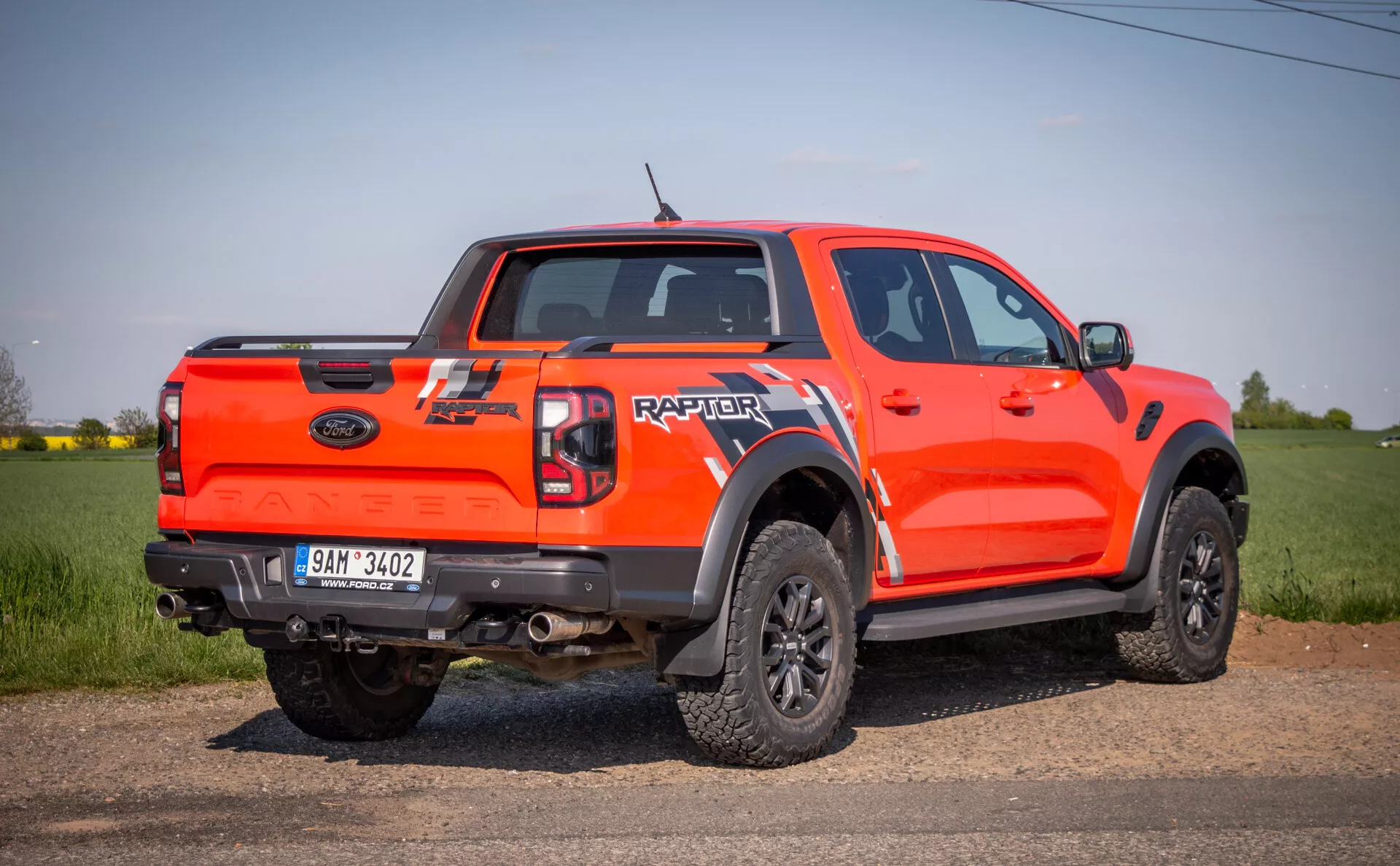Svetlo oranžový pickup Ford Raptor z roku 2022 z bočného zadného uhla, vonku na poli, ukazujúci jeho schopnosti pre jazdu v teréne s vysokou svetlou výškou, dverami double cab, veľkými čiernymi rozšíreniami podbehov, čiernymi kolesami s viacerými paprskami a terénnymi pneumatikami, a zadné označenie 'RAPTOR' na veku nákladového priestoru.