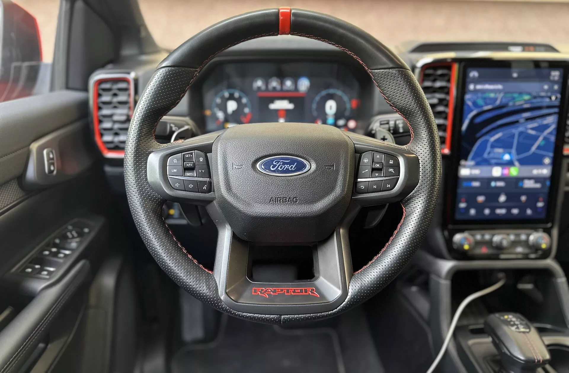 Interiér Ford Ranger Raptor 2022 s pohľadom spredu, ktorý zdôrazňuje kožený volant so troma ramenami a červeným prešívaním s logom Ford, digitálny prístrojový panel, moderný dotykový infotainment systém v režime navigácie, športovú palubnú dosku s červenými akcentmi a pokročilé pomôcky pre vodiča pre vysoko kvalitný, technologicky zameraný zážitok z jazdy