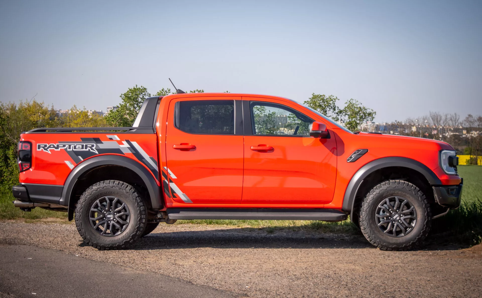 Ford Ranger Raptor 2022 v žiarivo oranžovej farbe, ukazuje agresívny štýl, schopnosti v teréne s veľkými čiernymi disky kolies a pneumatikami s hlbokým dezénom, zaparkovaný na spevnenej ploche vedľa trávy počas dňa