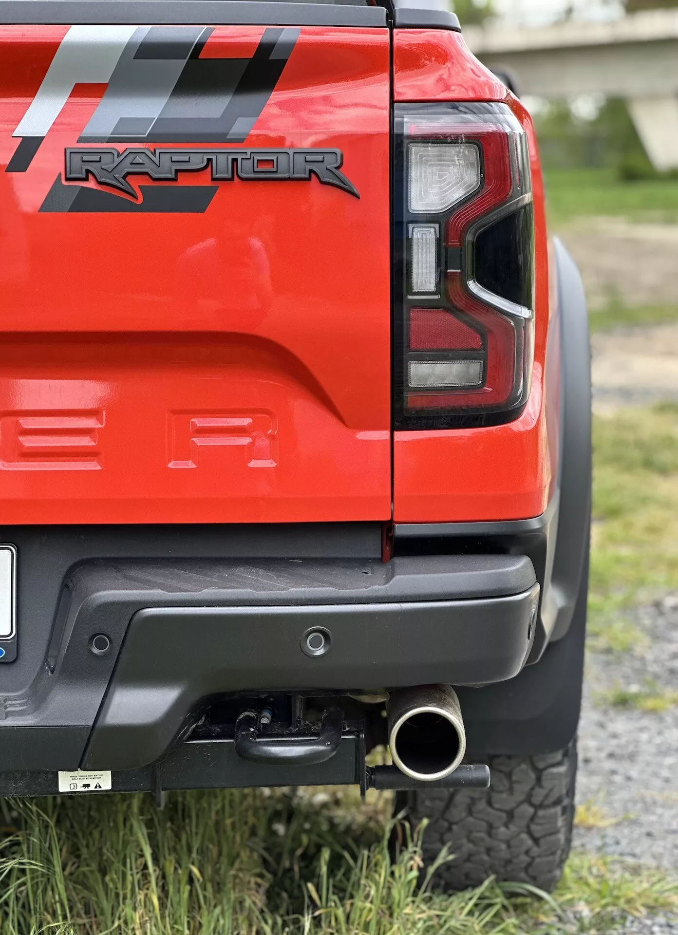 Detailný pohľad na zadný roh červeného Ford Ranger Raptor 2022 so zadným LED svetlom, čiernym zadným nárazníkom so sivým panelom, ťažným zariadením, leštenou koncovkou výfuku a terénnou pneumatikou, ktorý zdôrazňuje jeho schopnosti v teréne a dizajnové detaily.