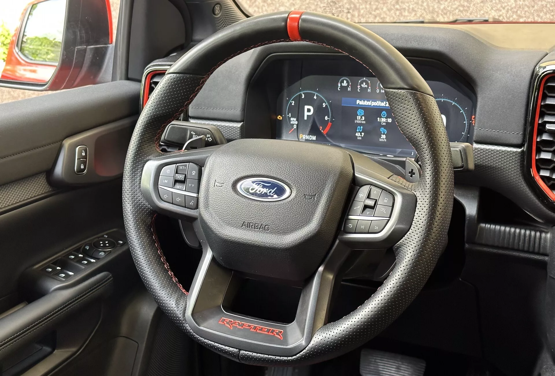 Detailný interiér Ford Ranger Raptor 2022 zameraný na volant pripomínajúci kožu s červeným prešívaním a multifunkčnými ovládacími prvkami a digitálny prístrojový panel zobrazujúci vozidlo v režime parkovania.
