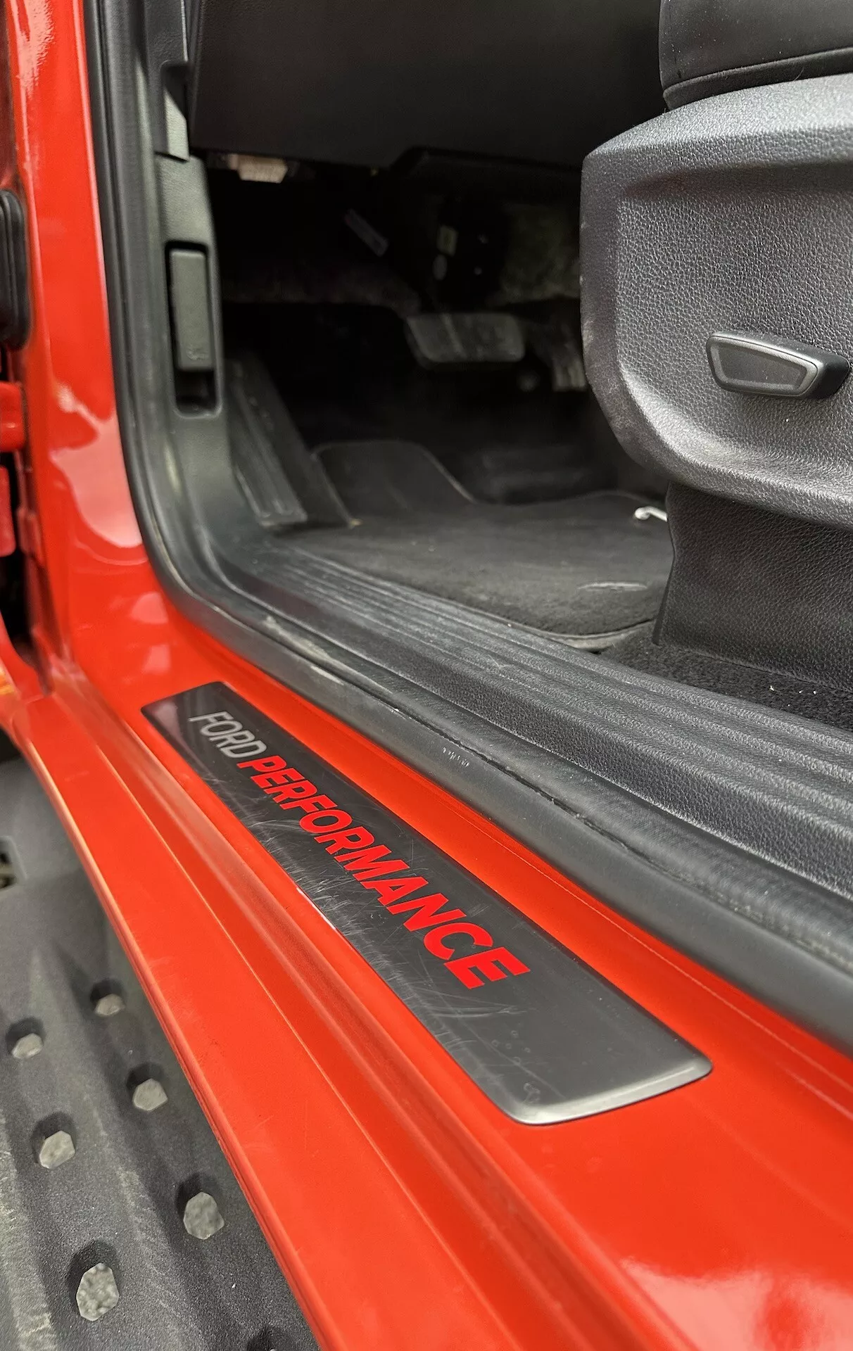 Detailný pohľad na prah dverí Ford Ranger 2022 s odznakom 'FORD PERFORMANCE', lesklý červený exteriér, čierny vnútorný koberec, šedé látkové sedadlo so športovým dizajnom a odolná čierna gumová podlahová rohož