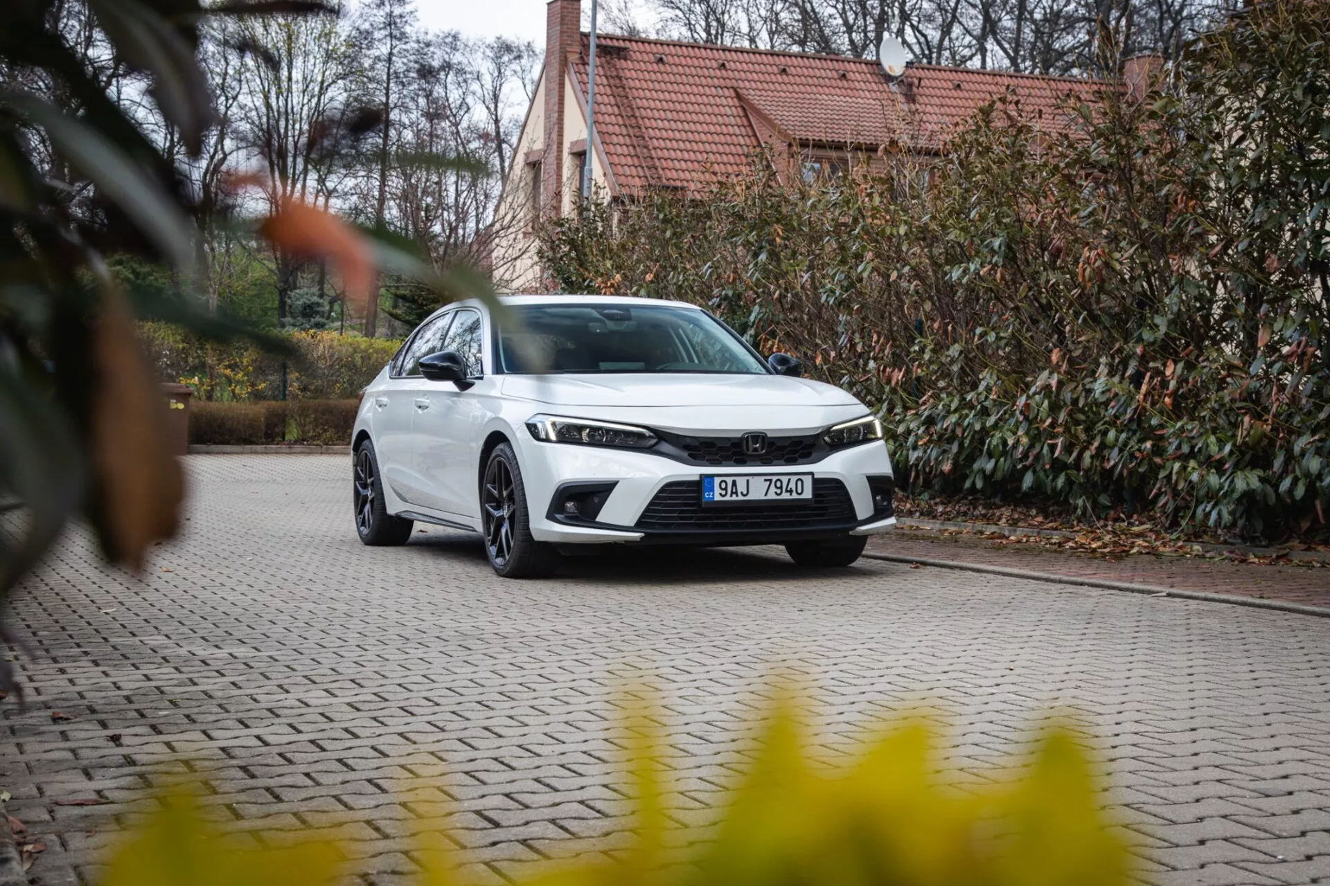 Moderný sedan Honda Civic 2022 zaparkovaný na dlažobnej príjazdovej ceste so záhradnou úpravou a budovou na pozadí, prezentujúci prednú mriežku, LED svetlomety a elegantný aerodynamický dizajn