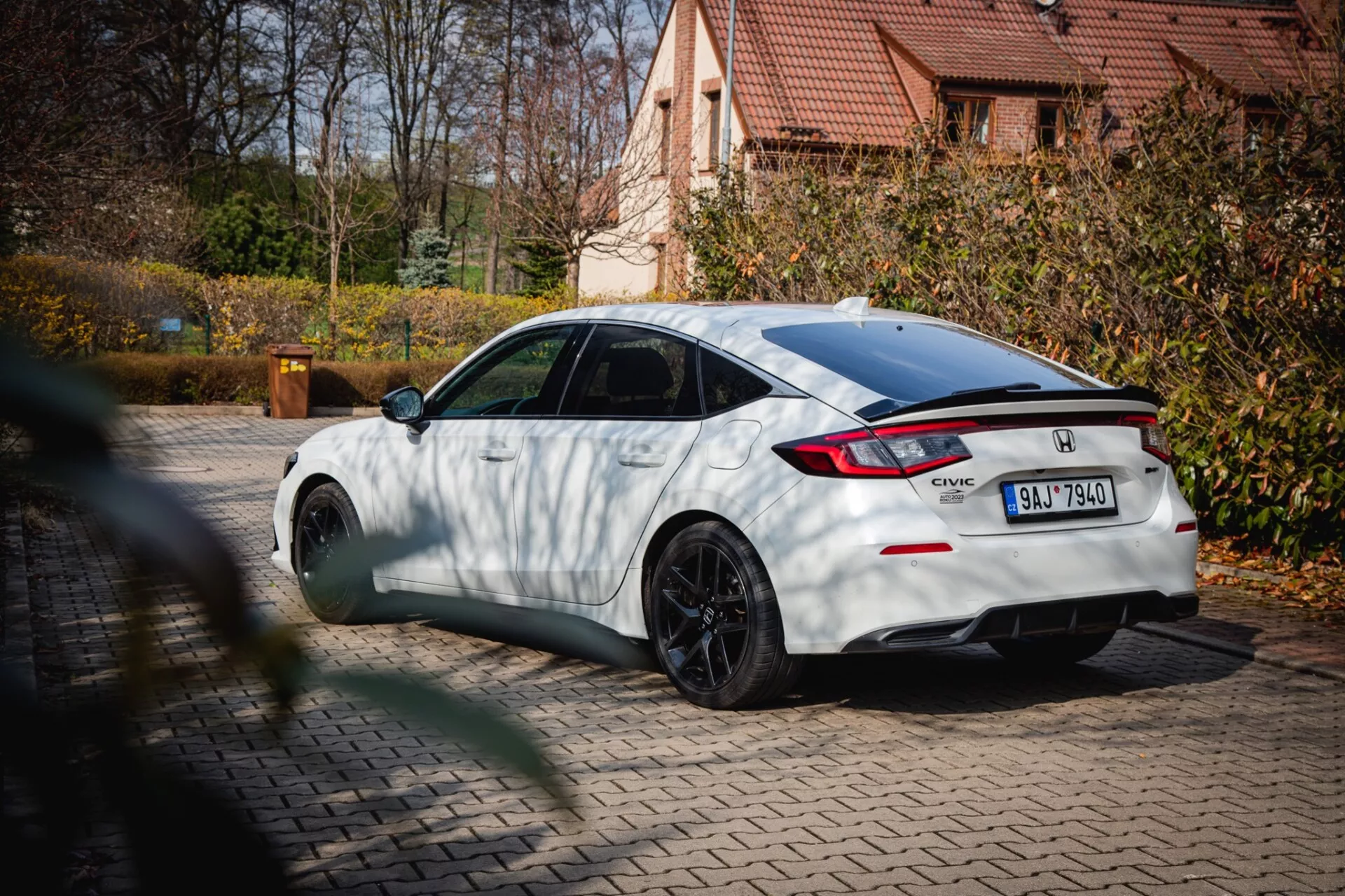 biely Honda Civic z roku 2022 s čiernymi viacramennými kolesami a zadným spojlerom, zaparkovaný na dlažbe s jesenným lístím a obytným domom na pozadí