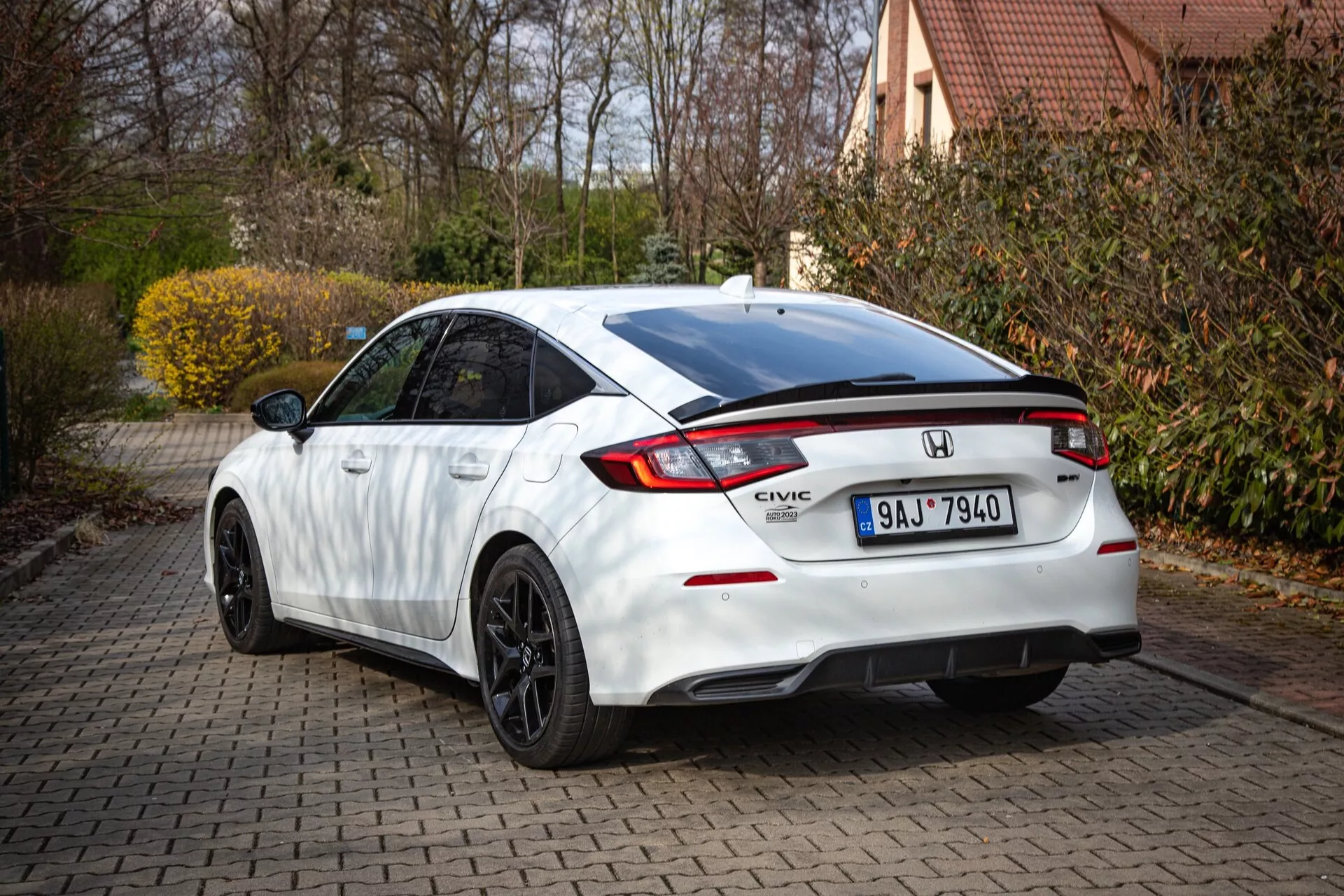 Biely Honda Civic 2022 zobrazujúci zadný a čiastočný bočný profil na tehlovej príjazdovej ceste, so zvažujúcou sa strechou, integrovaným spojlerom, LED brzdovým svetlom, charakteristickými zadnými svetlami a športovým nárazníkom s difúzorom