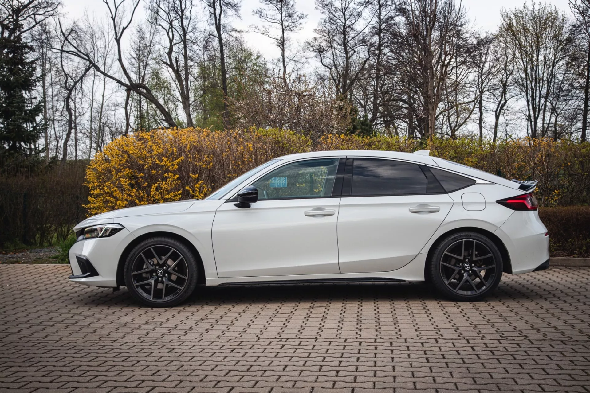 Biely štvordverový sedan Honda Civic z roku 2022 zaparkovaný na dlažbenej ploche s prepojenými tehličkami, zobrazený z bočného profilu so športovým dizajnom a modernými LED svetlometmi, zachytený z mierne zvýšeného uhlu na zvýraznenie plného bočného výhľadu vrátane tónovaných okien, športových zliatinových kolies a technologických prvkov, ako sú integrované smerové signály v bočných zrkadlách.
