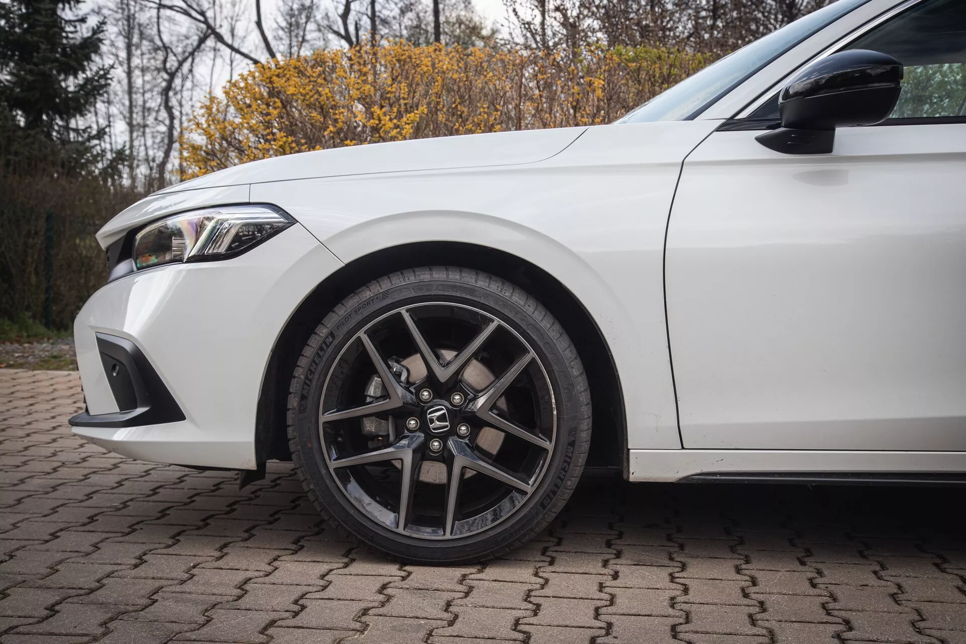 Biely Honda Civic 2022, zobrazuje predné ľavé koleso s dizajnom zliatiny s viacerými paprskami a nízkoprofilovou pneumatikou, spolu s detailmi blatníka a časti predného nárazníka na tehlovej ploche