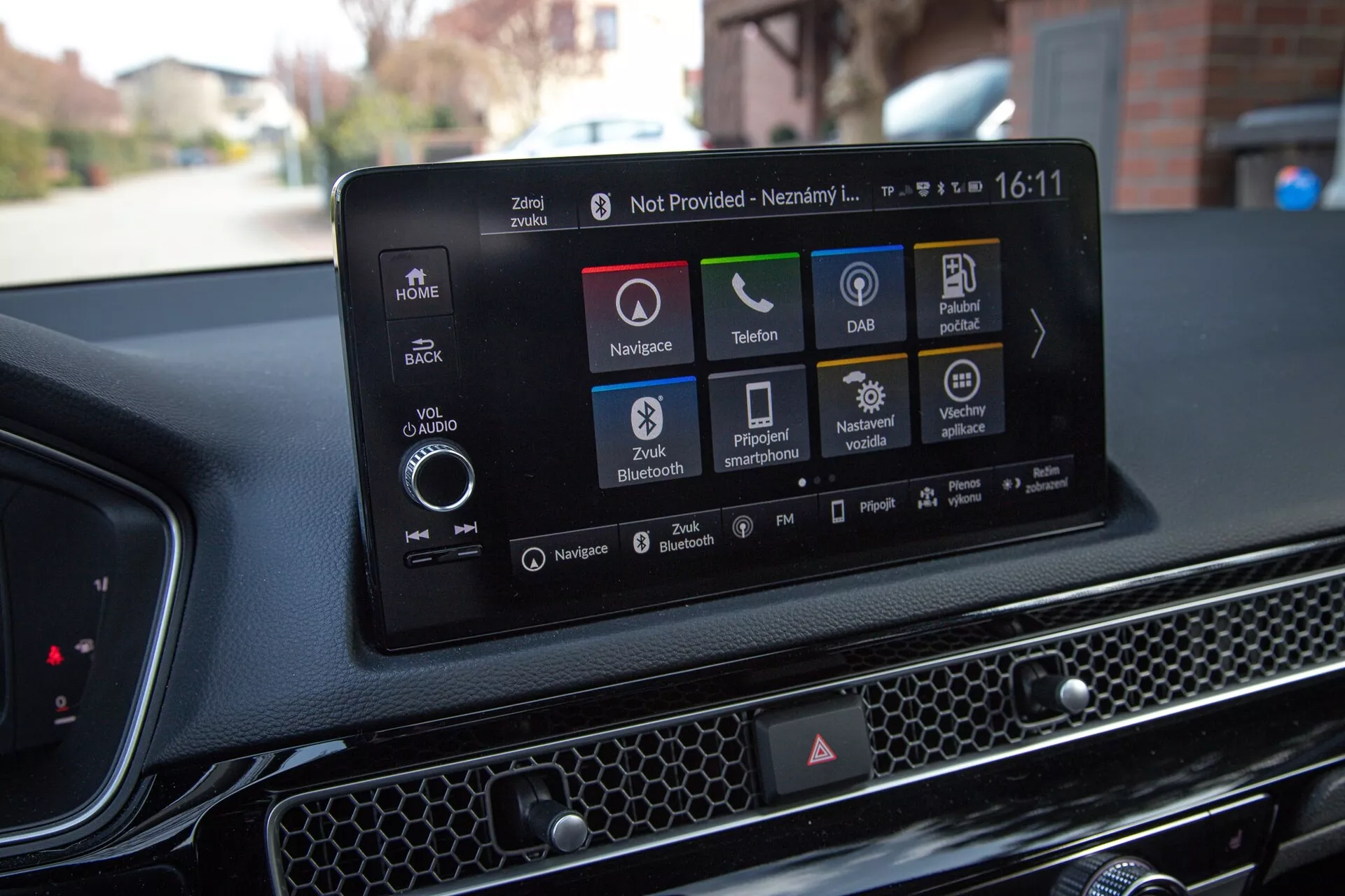 Interiér Honda Civic 2022 zobrazujúci moderný dotykový informačný systém s navigáciou, ikonami pre prehrávanie zvuku a médií, pripojenie telefónu, DAB, informácie o stave vozidla, Bluetooth a možnosti nastavenia vozidla, proti texturovanej palubnej doske s včelou plástvou.