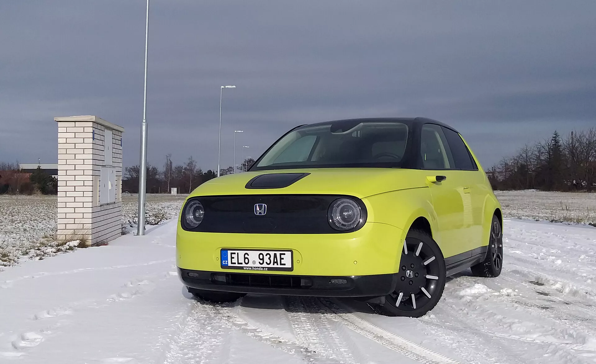 Honda e 2020 vo svetložltej farbe na povrchu pokrytom snehom s zachmúrenou oblohou, s LED svetlomety, technológiou elektrického vozidla s pokročilými asistenčnými systémami, čiernymi zliatinovými kolesami a zimnými pneumatikami.