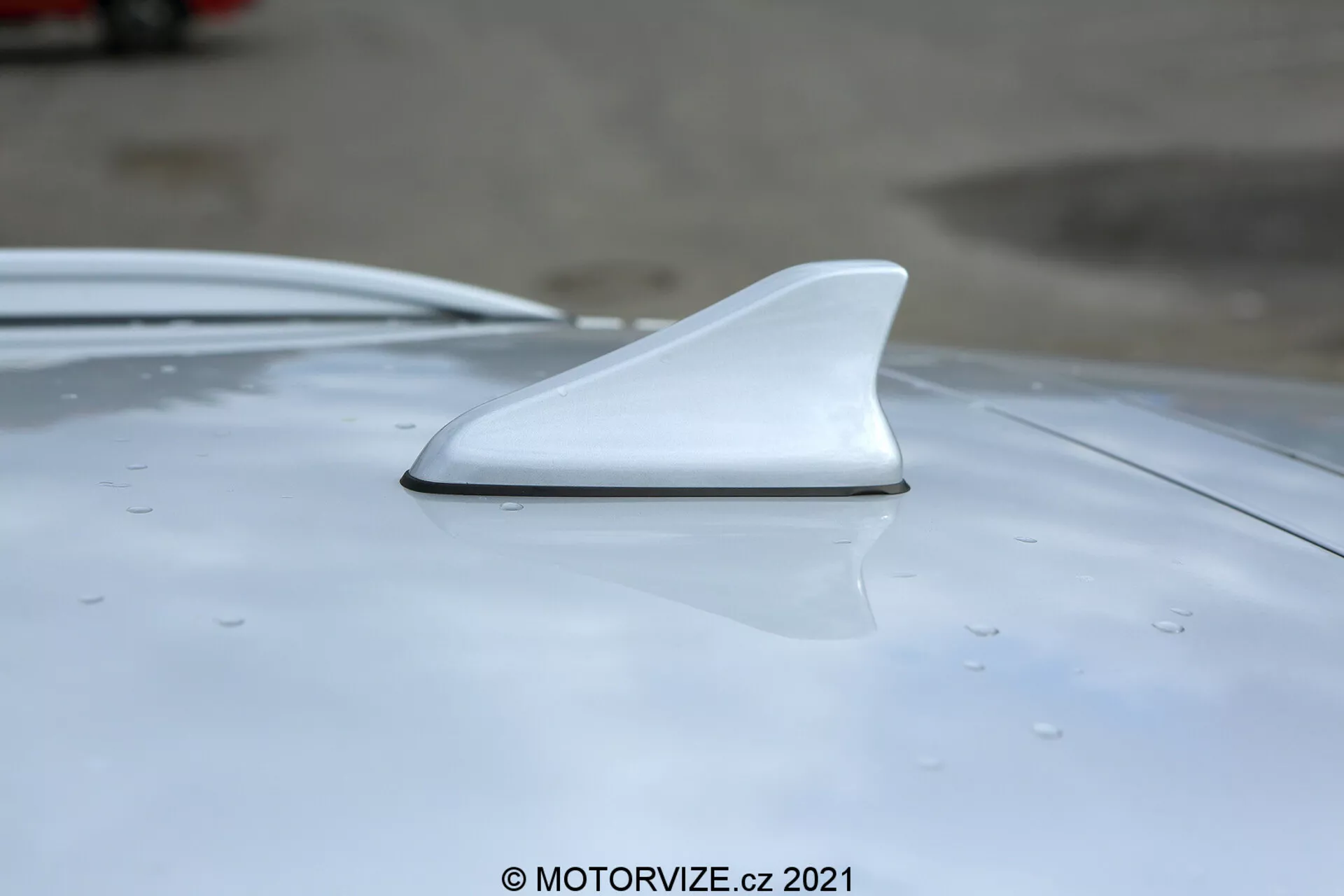 Detailný záber na bielu anténu v tvare žraločej plutvy na streche vozidla Hyundai i30 2020, ukazuje jej aerodynamický dizajn z bočného a mierne zvýšeného uhla so zameraním na hladkú integráciu a kvapky vody naznačujúce nedávny dážď alebo umytie.