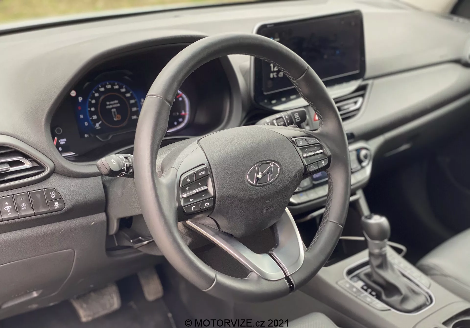 Pohľad na interiér Hyundai i30 Kombi 2020 zameraný na volant a palubnú dosku. Fotografia je zhotovená z miesta spolujazdca a ukazuje elegantný volant s ovládacími tlačidlami, digitálny prístrojový panel a infotainment systém. Moderný dizajn obsahuje kožený volant so strieborným spodným lúčom, vetracie otvory a minimálne fyzické ovládače, zdôrazňujúc pokročilé automobilové technológie a ergonomický dizajn.