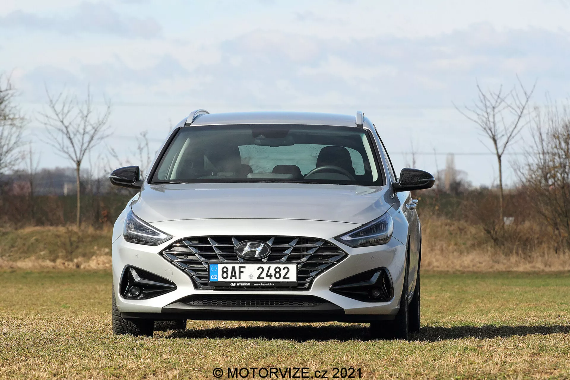 Predný uhol pohľadu na Hyundai i30 2020 Kombi zobrazujúci polygonálnu mriežku s logom výrobcu, ostré hranaté svetlomety, tvarovaný predný nárazník, jemné línie kapoty a bočné zrkadlá z trávnatej oblasti a prírodného pozadia.