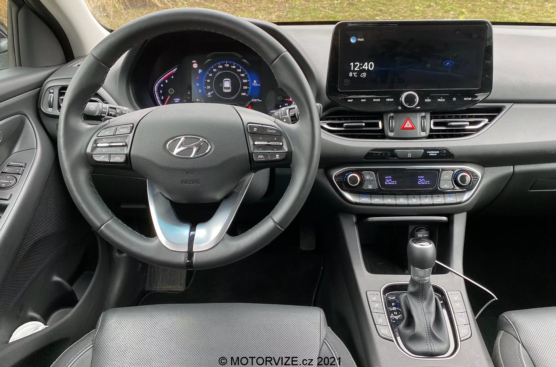 Pohľad na interiér Hyundai i30 Kombi 2020 z miesta vodiča, s modernou prístrojovou doskou a volantom s logom Hyundai, ktorý má ovládacie tlačidlá pre funkcie ako tempomat, audio a hands-free volanie. Digitálny prístrojový panel zobrazuje rýchlosť a ďalšie informácie o vozidle. Na stredovej konzole je veľká dotyková obrazovka infotainmentu pre navigáciu a médiá, vedľa sú fyzické ovládacie prvky klimatizácie, čo naznačuje dvojzónový klimatizačný systém. Stredová konzola obsahuje radiacu páku s koženým poťahom a USB port, čo zdôrazňuje pokročilé pripojenie a technologické vybavenie.